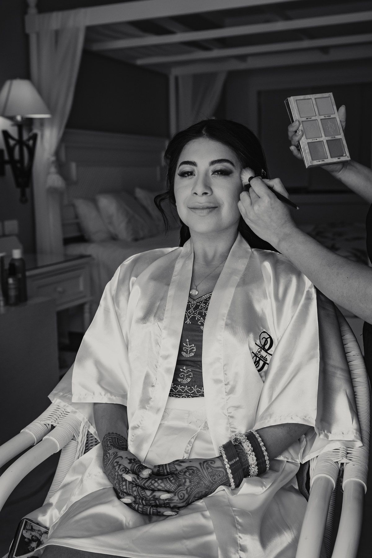 Bride in satin robe with henna hands gets makeup applied by artist in black and white wedding preparation photo.