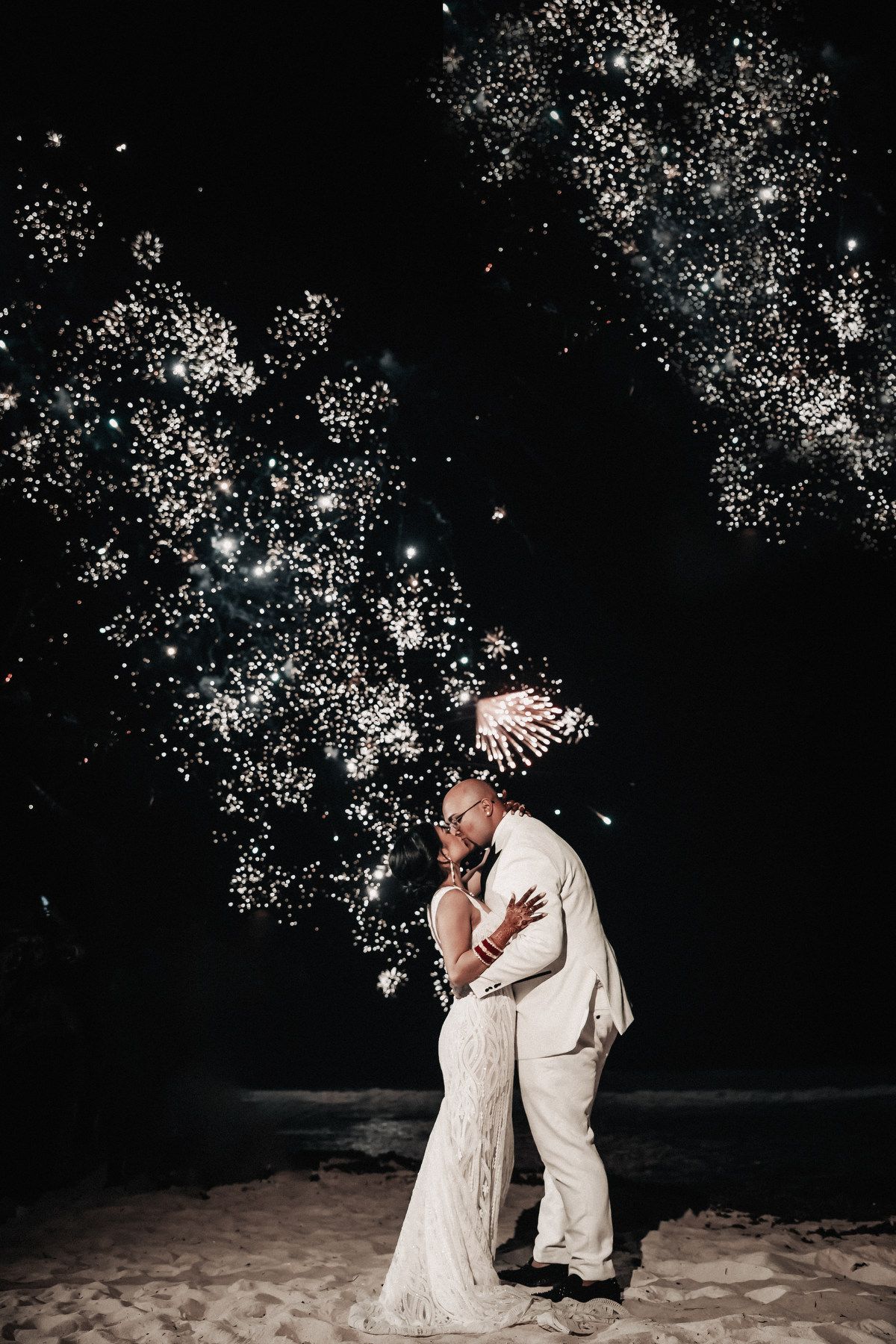 Bride and groom kiss on beach under spectacular fireworks display at night wedding celebration.