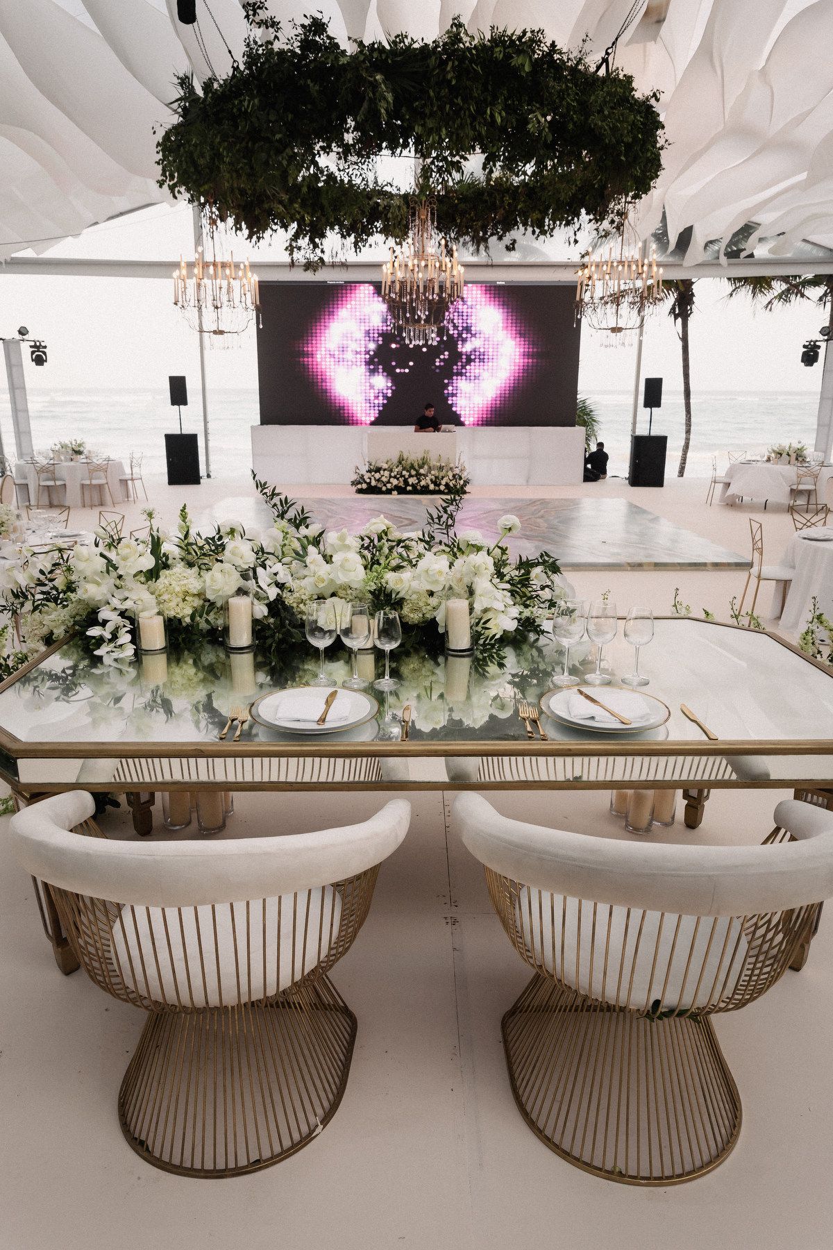 Elegant beach wedding reception with sweetheart table, white floral centerpiece, and circular greenery chandelier overhead.