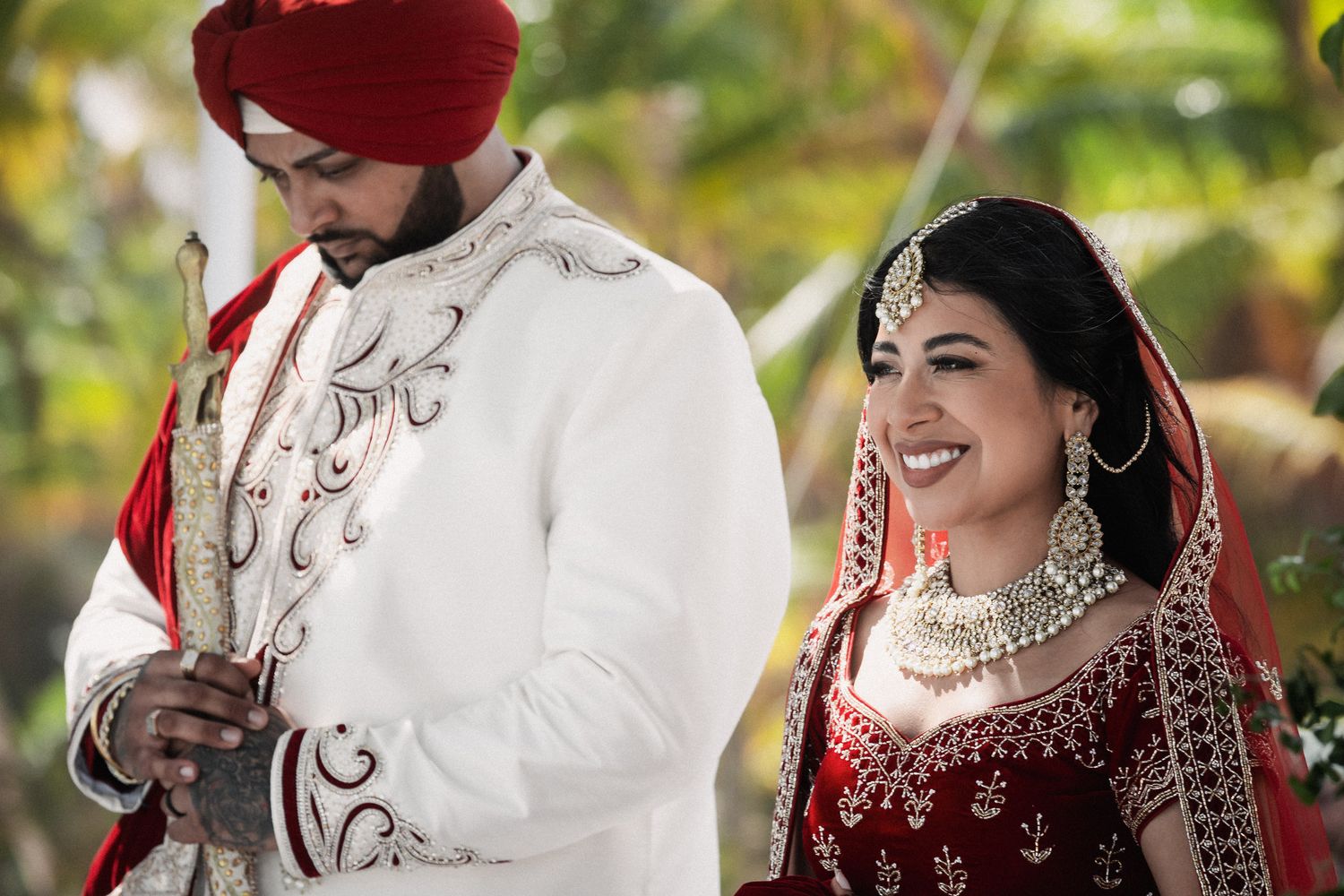 Sikh bride and groom smile together in traditional red and white wedding attire with jewelry.