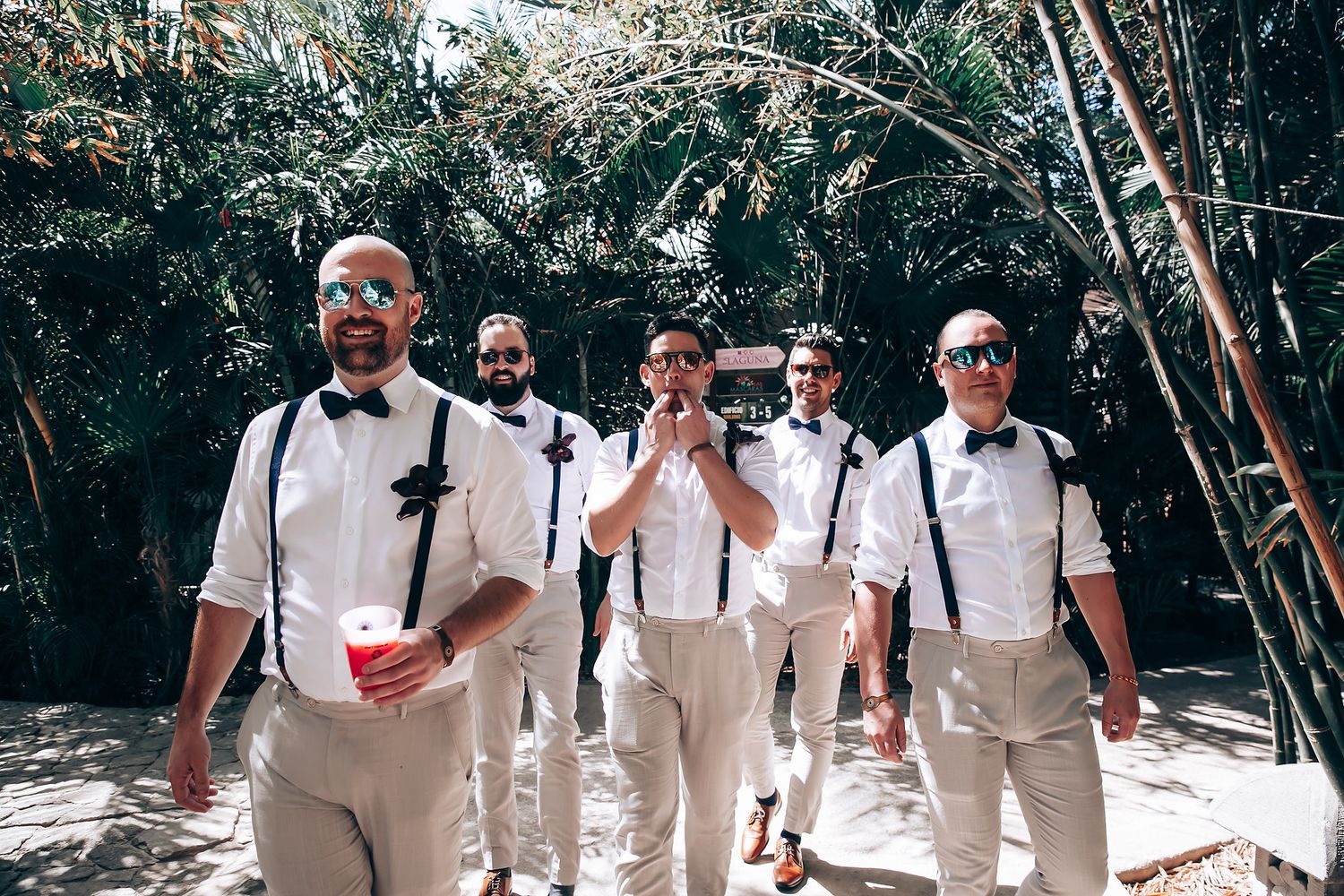 Wedding party members in light colored attire walk along a shaded outdoor path.