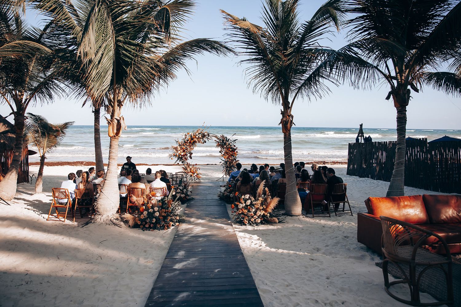 Palm trees line a tropical beach wedding ceremony setup with orange floral decorations at sunset.