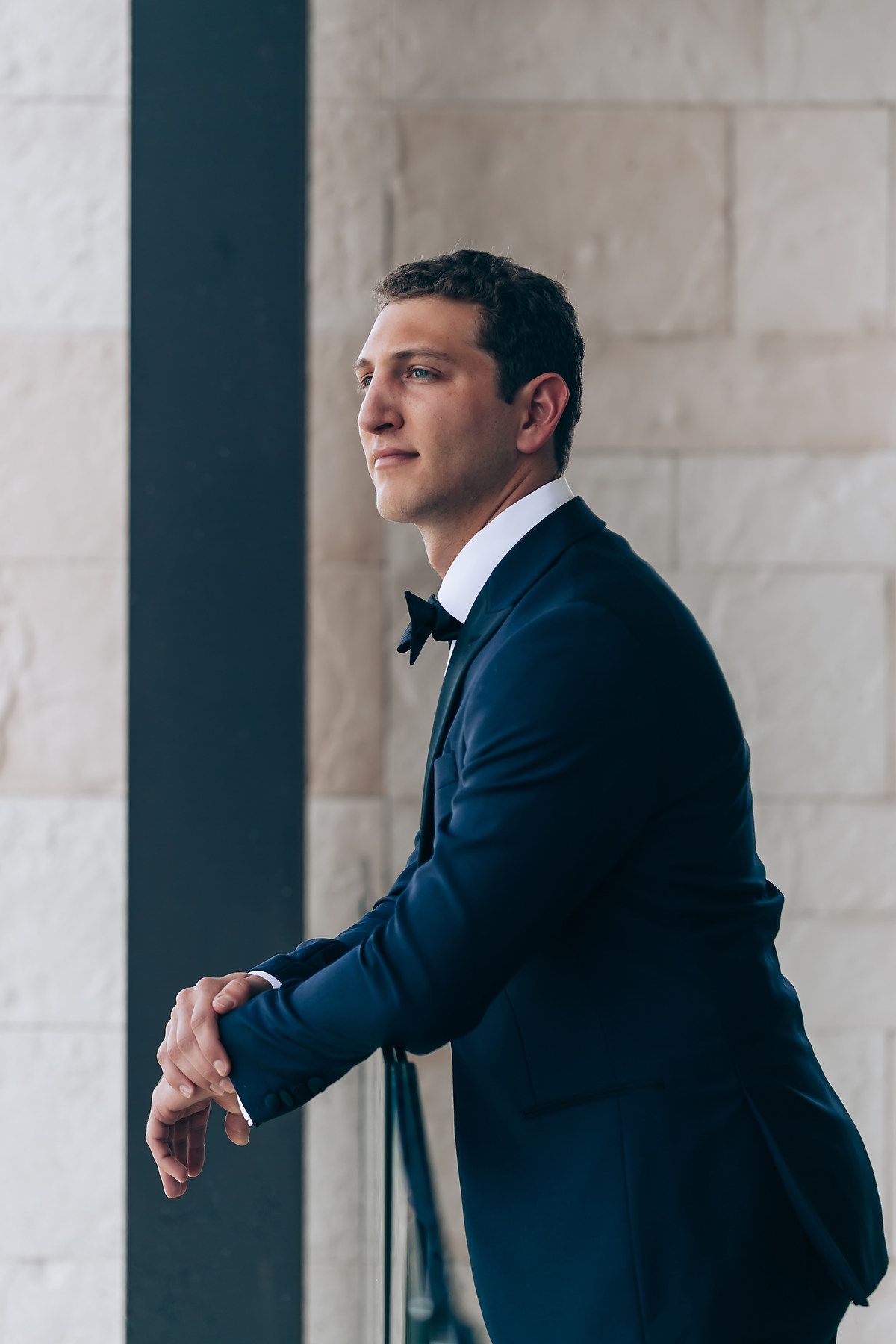 Man in navy suit and bowtie standing against white columned architecture.