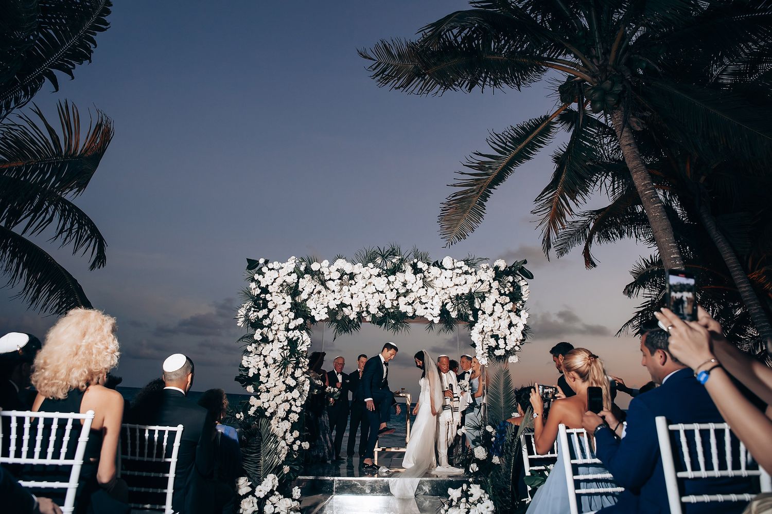 Outdoor wedding reception under palm trees with white floral decorations at sunset.