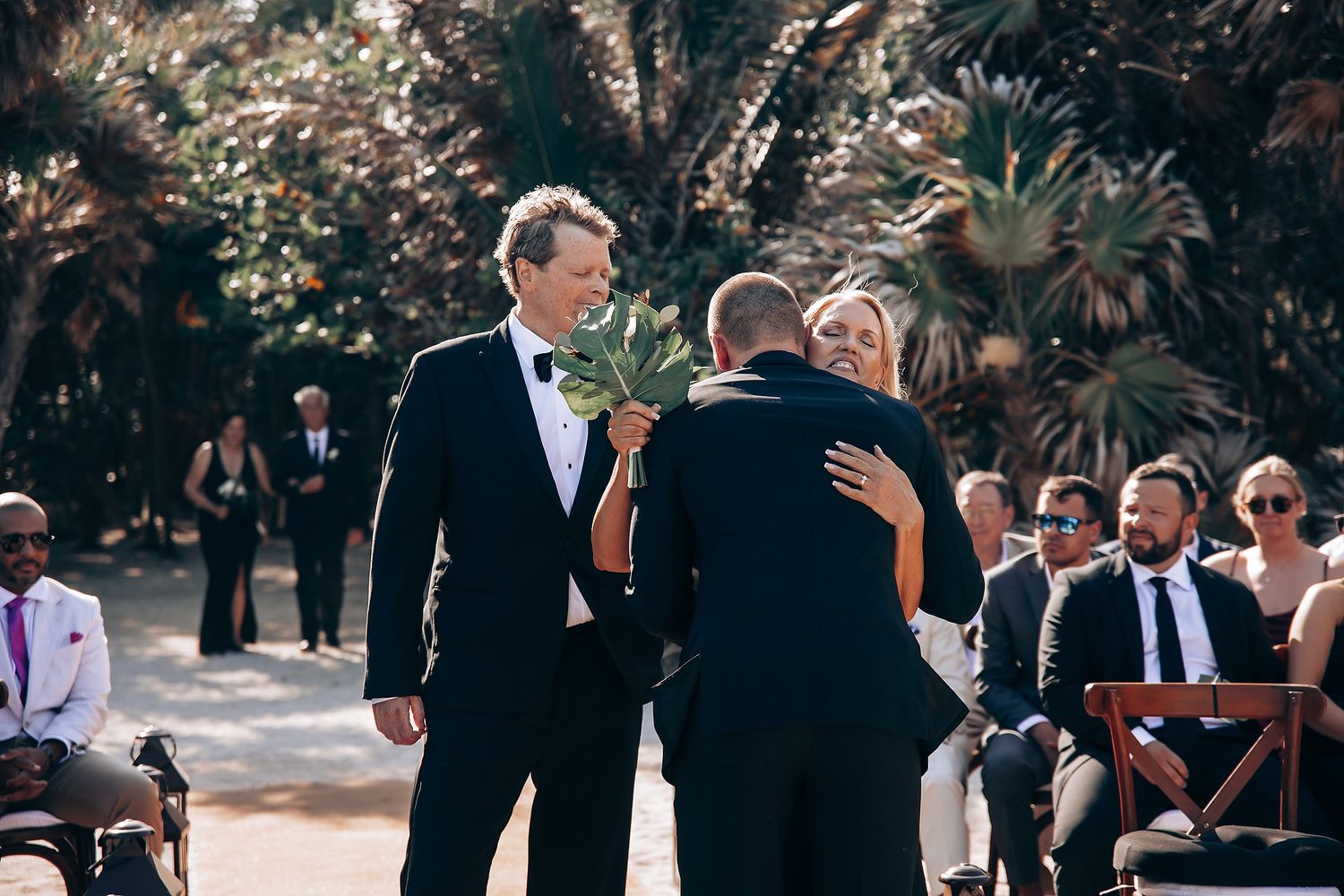 Wedding ceremony guests gathered outdoors under natural tree canopy.