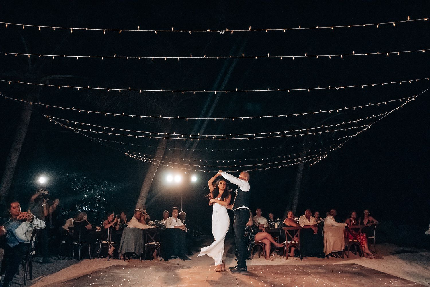 Wedding reception at night with string lights overhead and guests seated at tables under the stars.