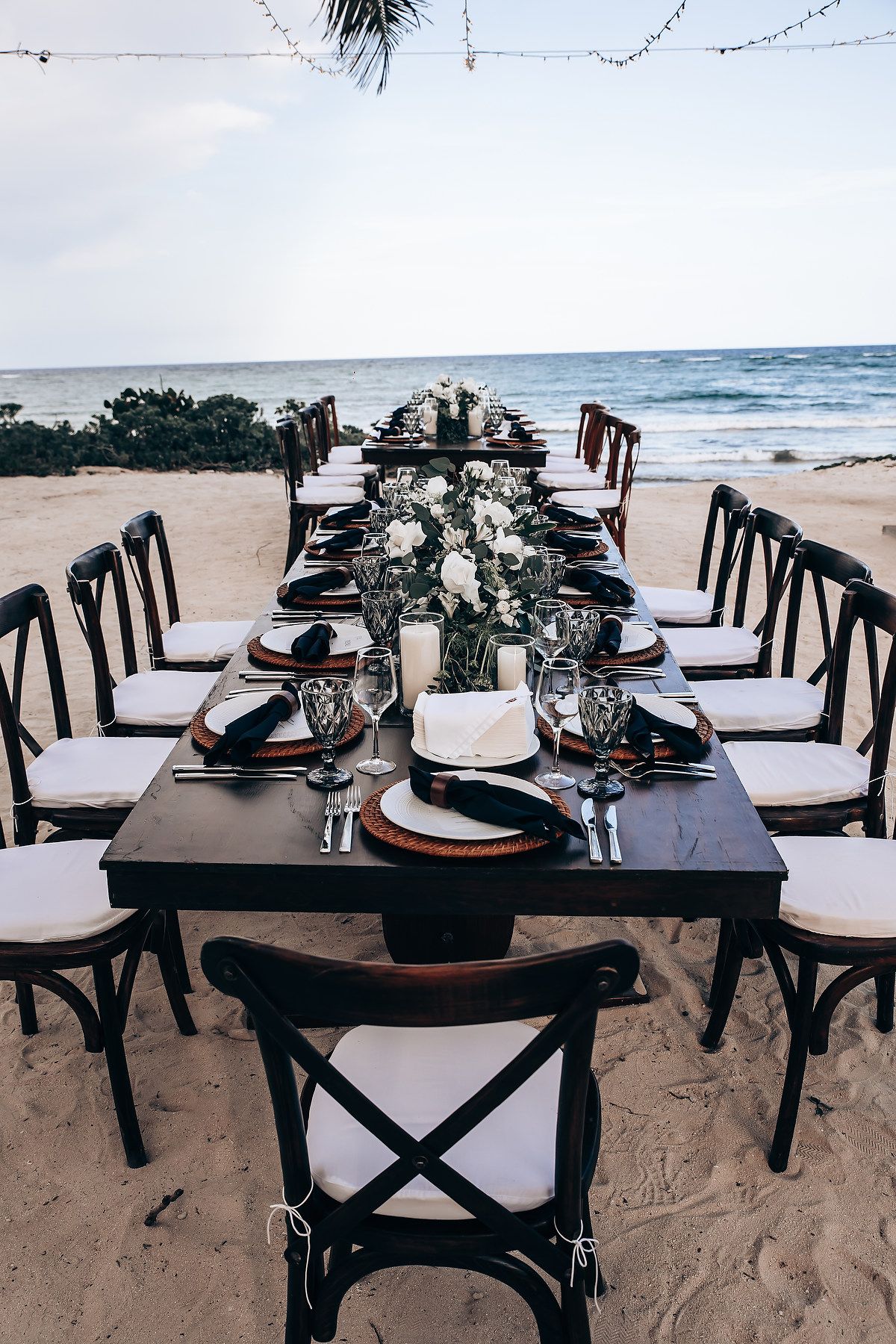 Beach wedding reception setup with dark wooden tables and chairs arranged on the sand.