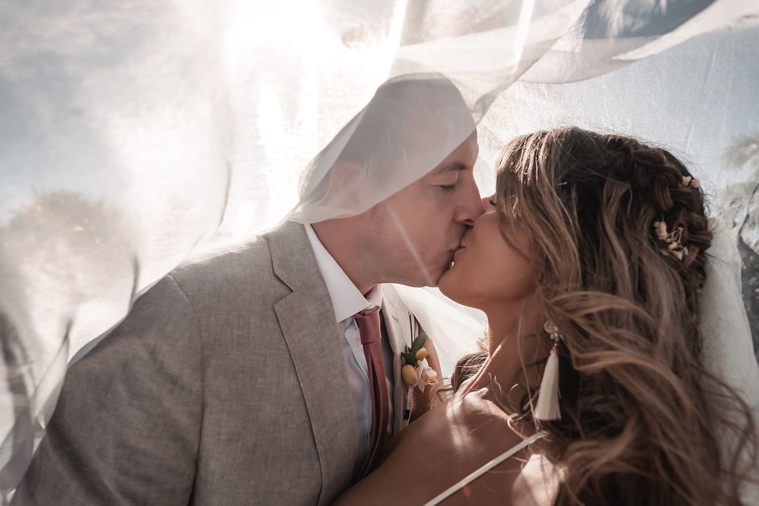 Bride and groom sharing a romantic kiss under a flowing white veil on their wedding day.