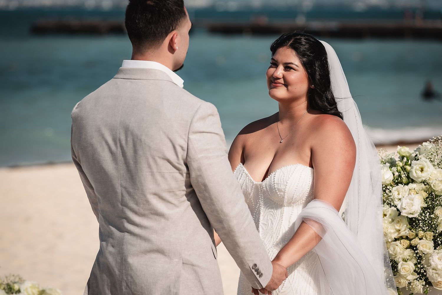 Bride in strapless white gown and veil holds hands with groom in beige suit during beach wedding ceremony.