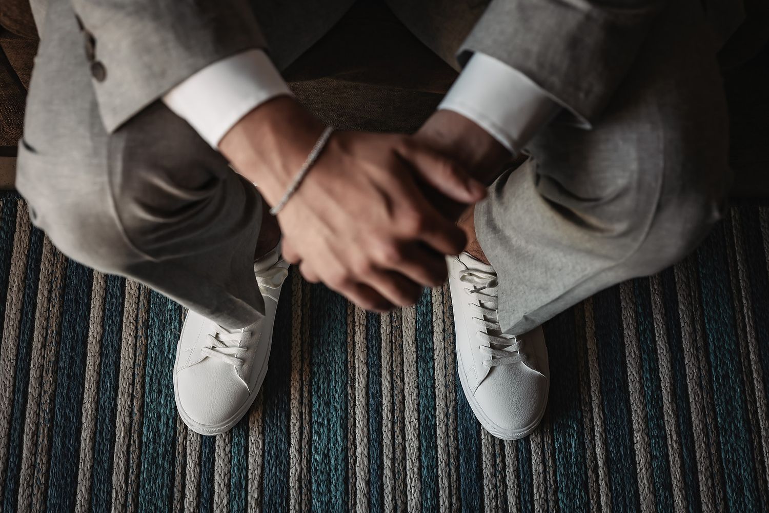 Man in gray suit and white leather sneakers sitting with hands clasped on striped carpet floor.