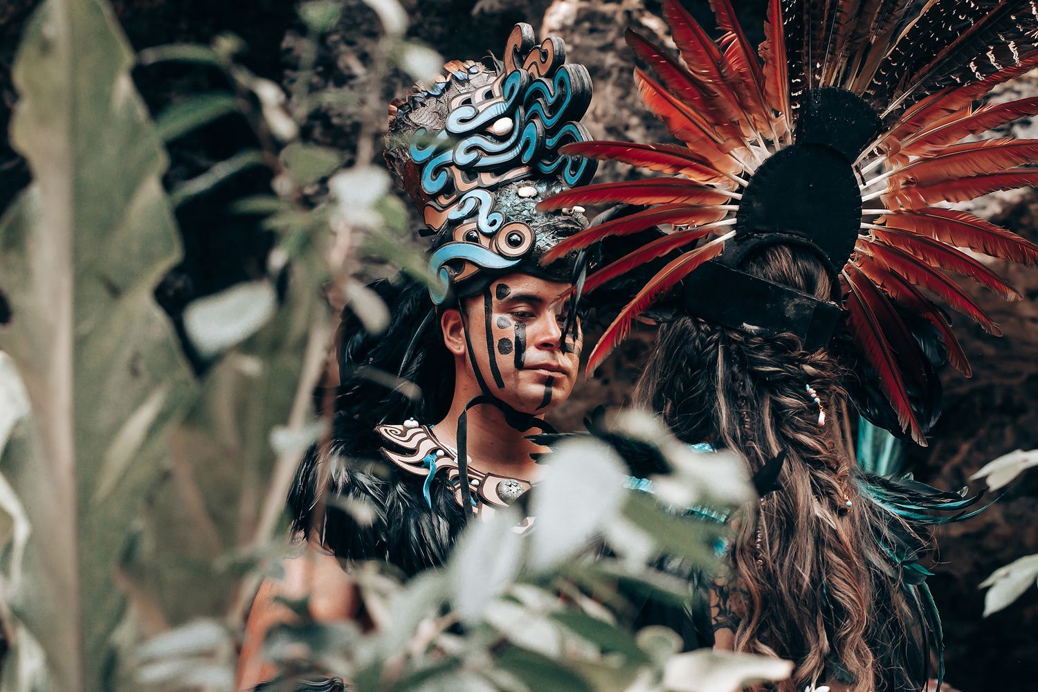 Performer in vibrant red and blue feathered Aztec headdress with face paint stands among tropical foliage.