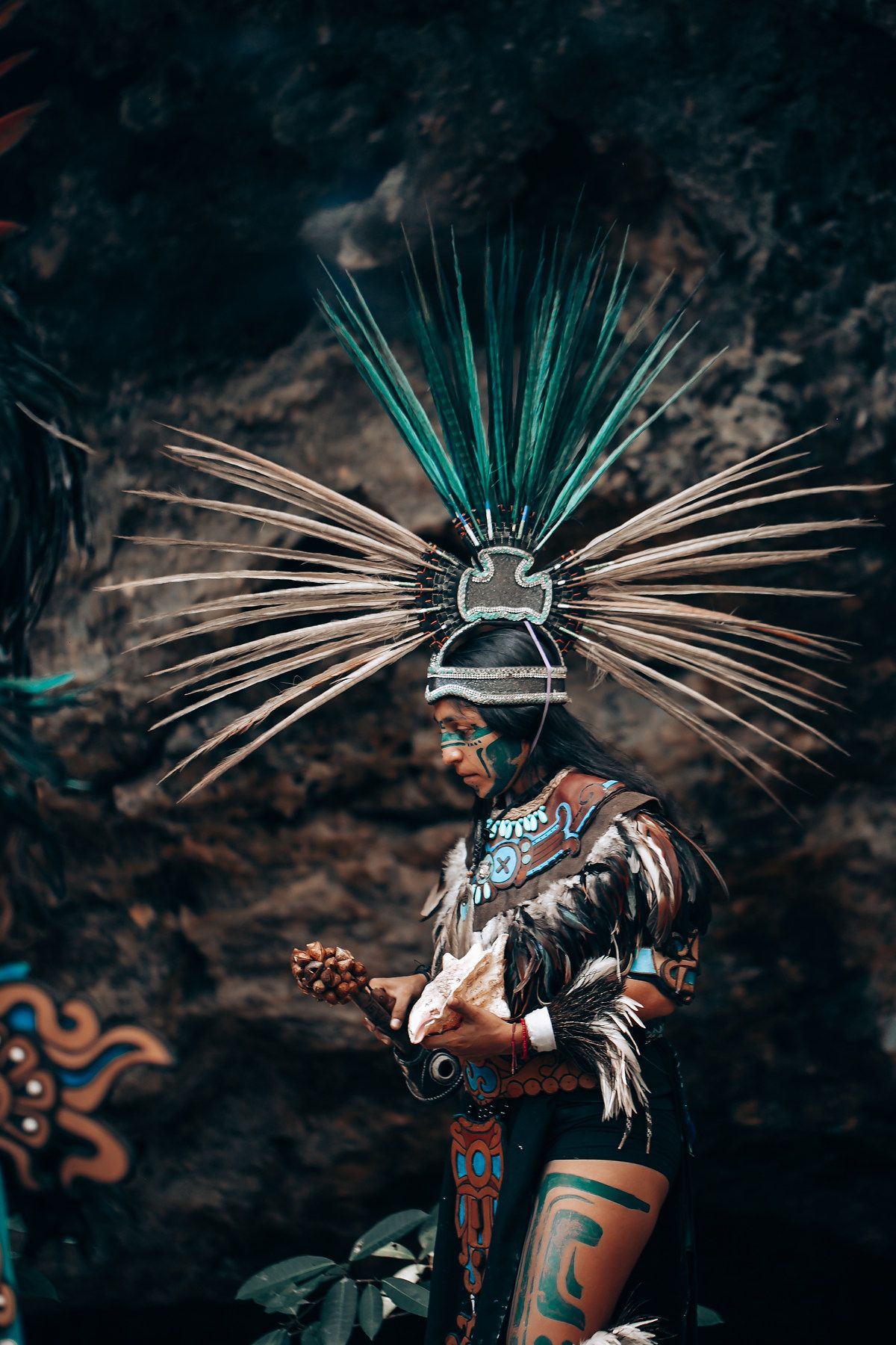 Mayan warrior performer in elaborate feathered headdress and traditional costume holding a ritual instrument outdoors.