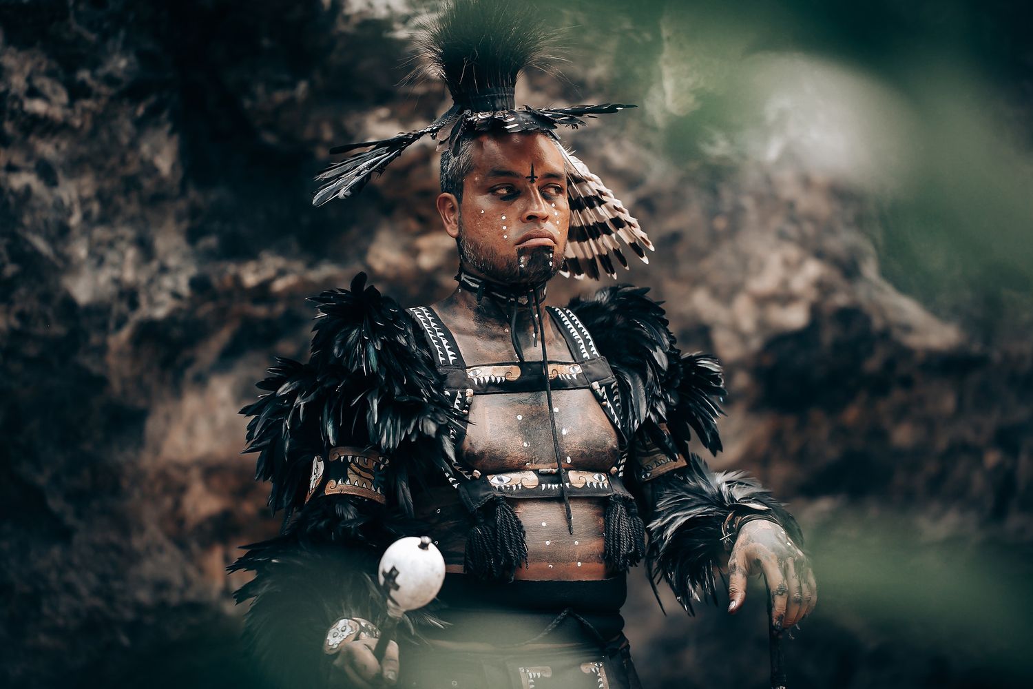 Person in elaborate black feathered indigenous ceremonial costume and headdress seated outdoors.