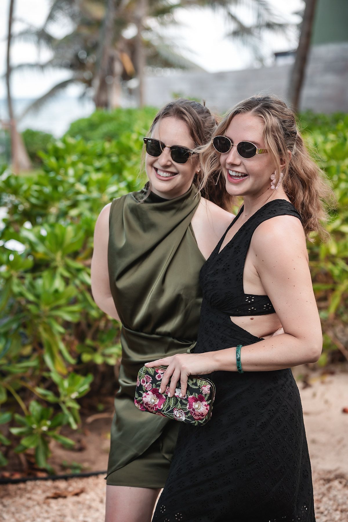 Two smiling women in elegant green and black dresses wearing sunglasses at a tropical outdoor wedding venue.