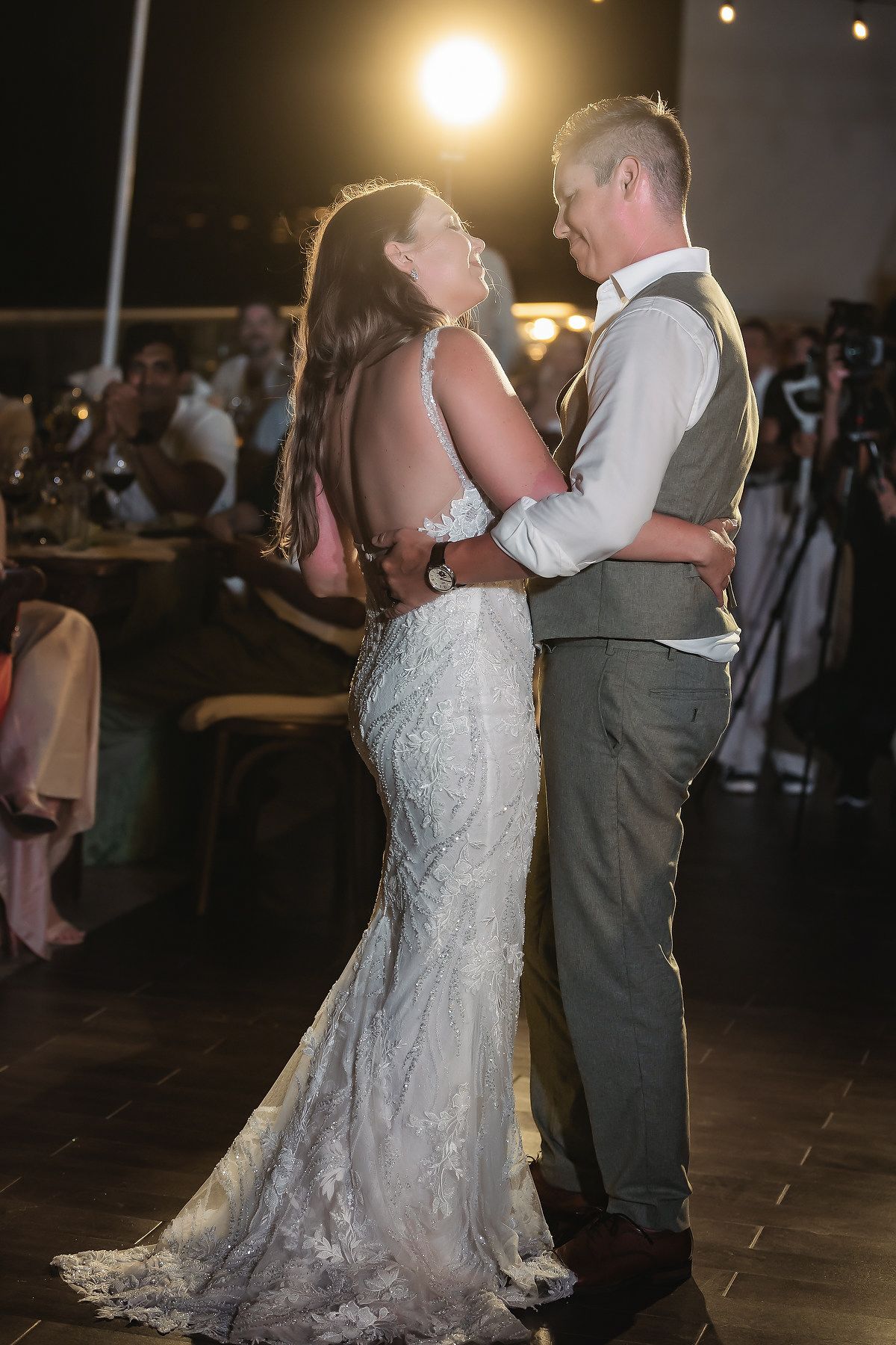 Bride and groom share their first dance at wedding reception, bride in lace gown, groom in gray vest and trousers.