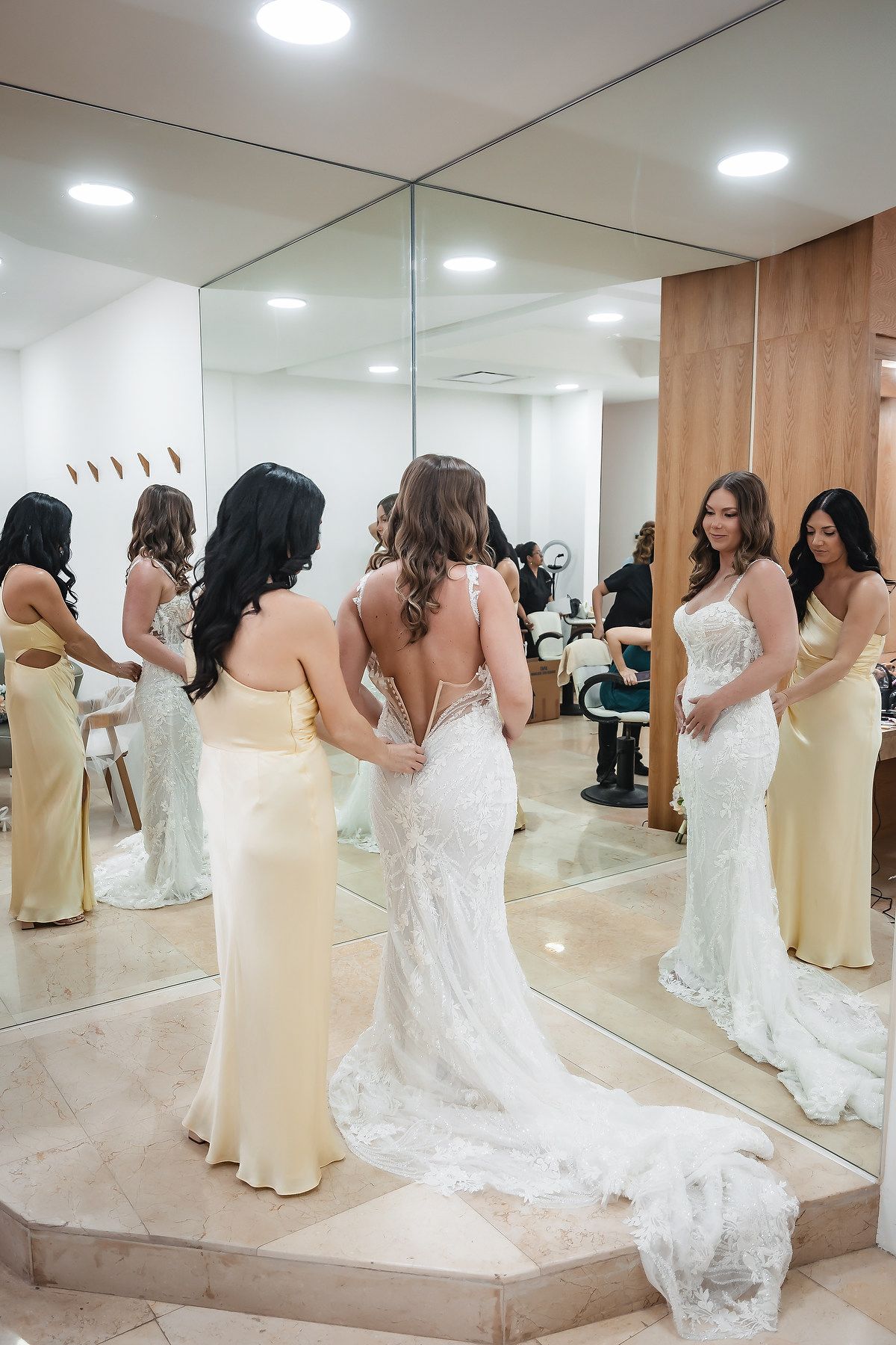 Bride in lace gown with bridesmaids in yellow dresses helping her prepare in a mirrored bridal suite.