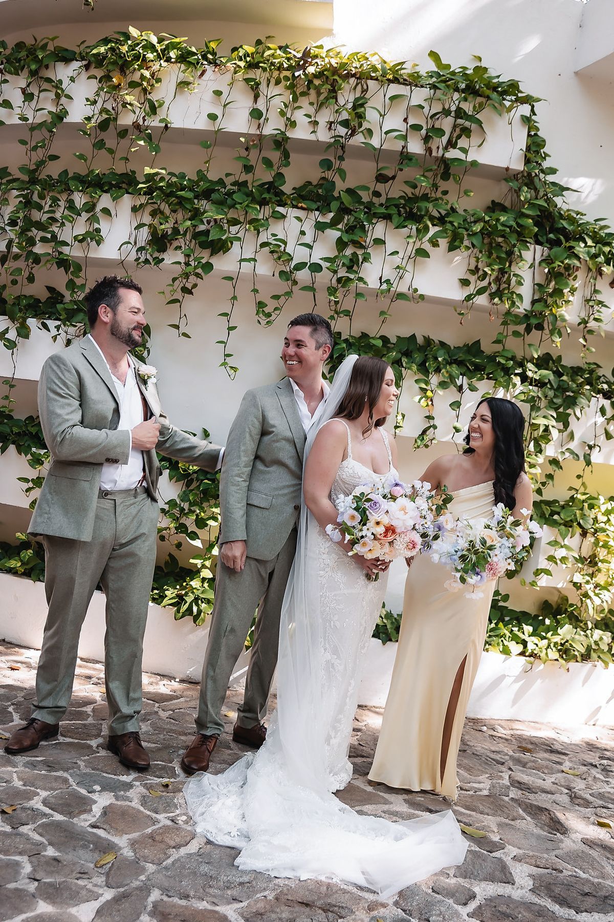 Wedding party laughing together outdoors under lush green ivy wall, bride holding colorful bouquet in elegant white gown.