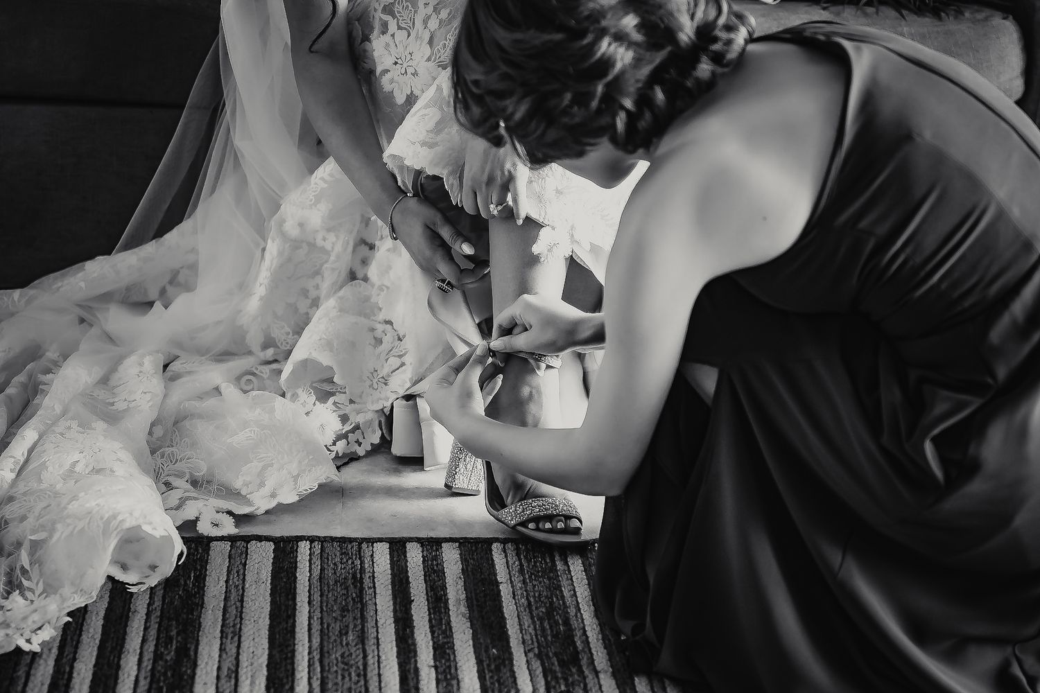 Bridesmaid kneeling to help bride with wedding shoes in elegant black and white photo.