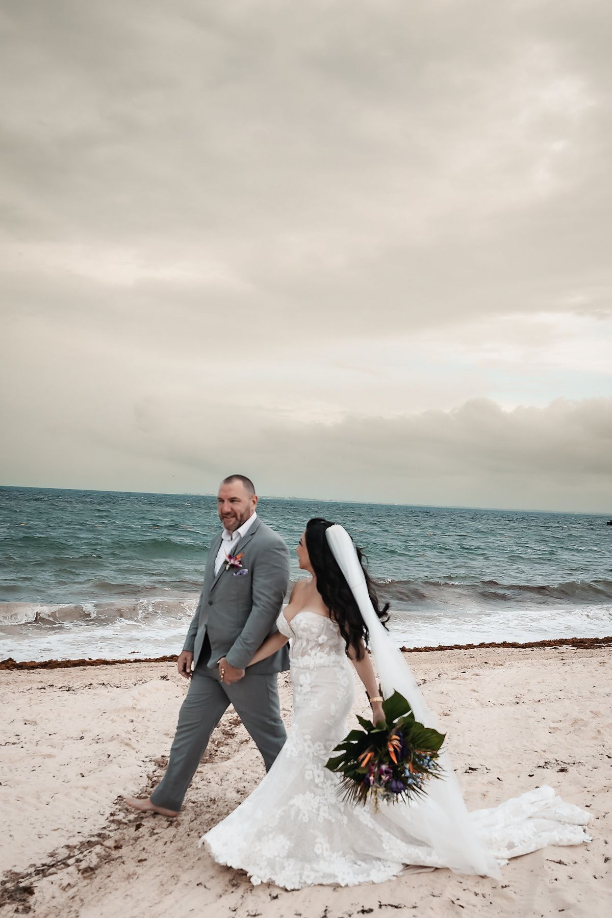 Bride and groom walk hand-in-hand on a sandy beach, she in a lace gown with veil, he in a gray suit.
