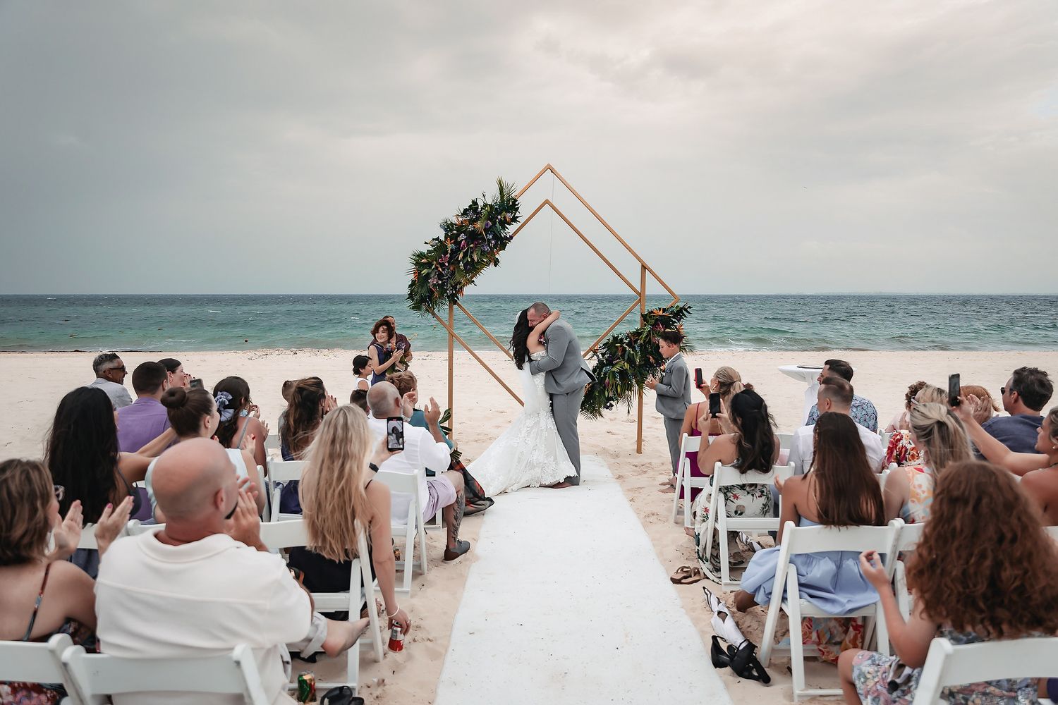 Romantic beach wedding ceremony with couple at geometric floral arch, guests seated on white chairs by the ocean.