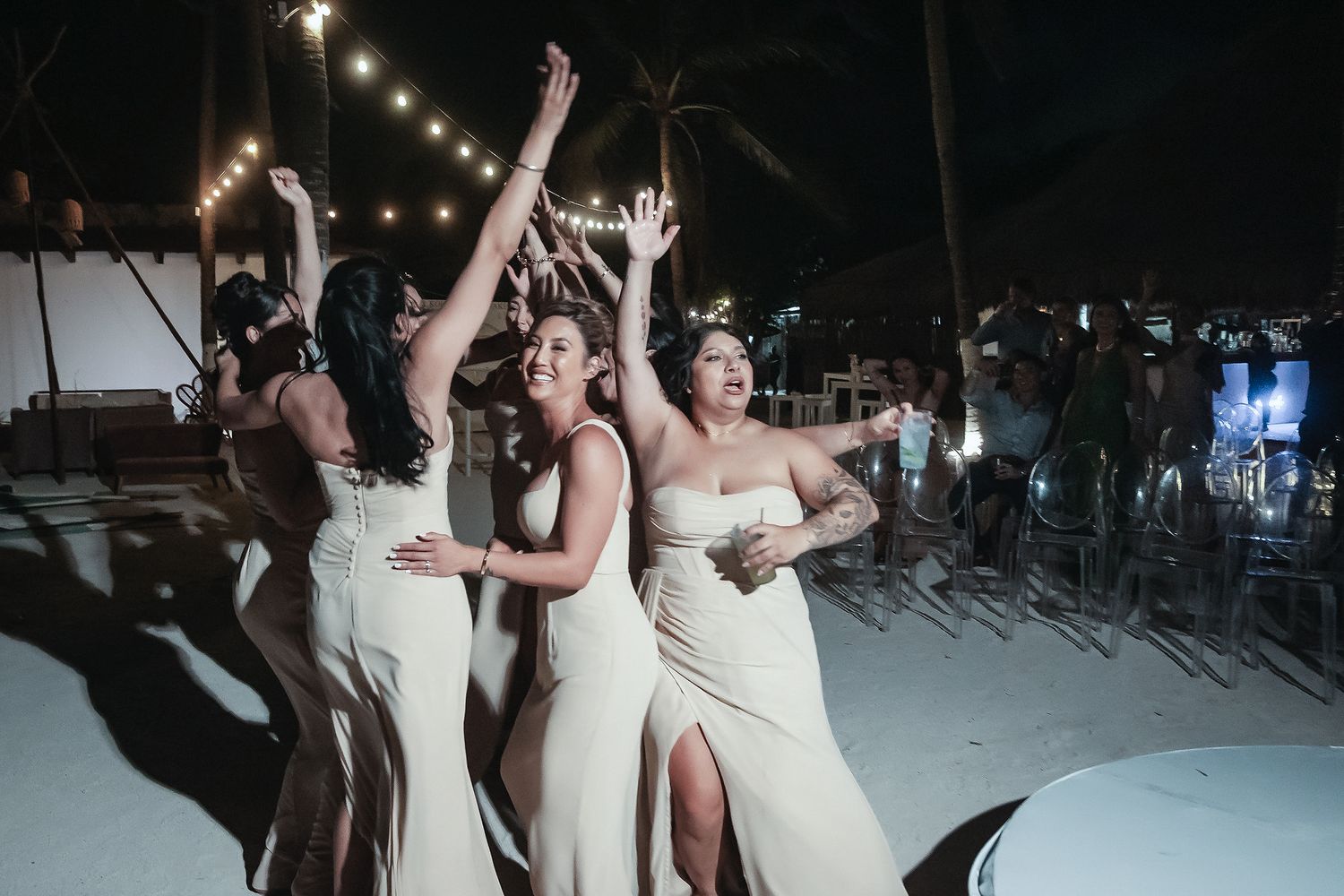 Bridesmaids in white dresses dancing joyfully at an outdoor beach wedding reception at night with string lights.