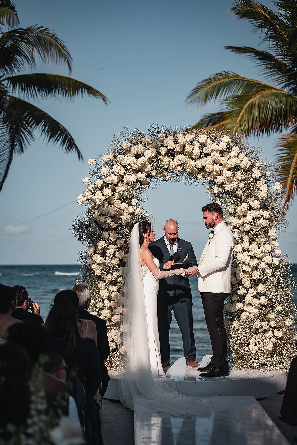 Couple exchanges vows under a floral arch adorned with white roses and baby's breath at a beachside wedding ceremony.