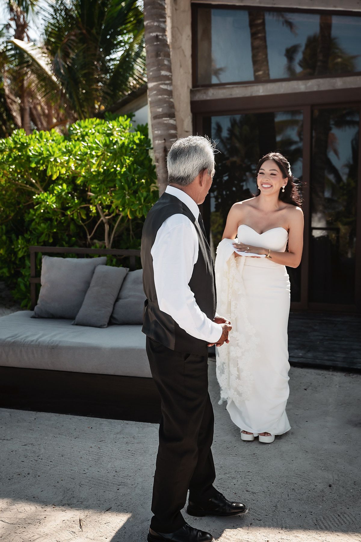 A smiling bride in a white strapless gown shares a first look moment with an older man in a black suit outdoors.