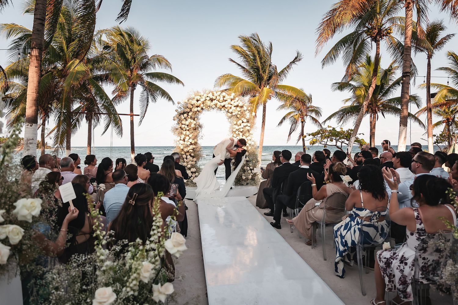 Romantic beachside wedding ceremony with floral arch, white aisle, palm trees, and ocean backdrop at sunset.