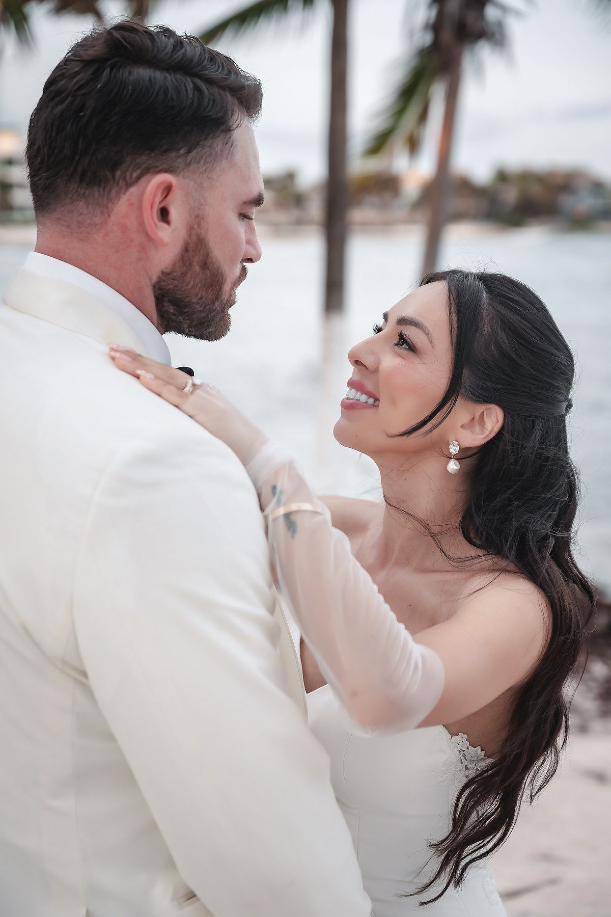 Bride in white dress smiles at groom in white suit during beach wedding photo session with palm trees.