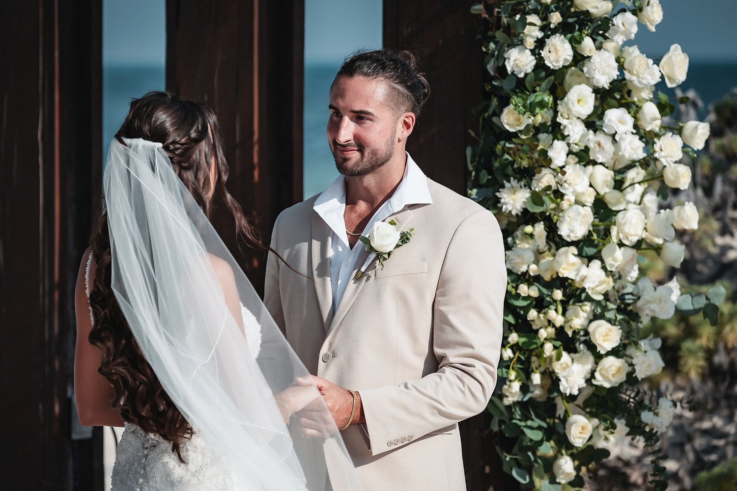 Groom in beige suit smiling at bride in white veil during outdoor wedding ceremony with white floral arch.