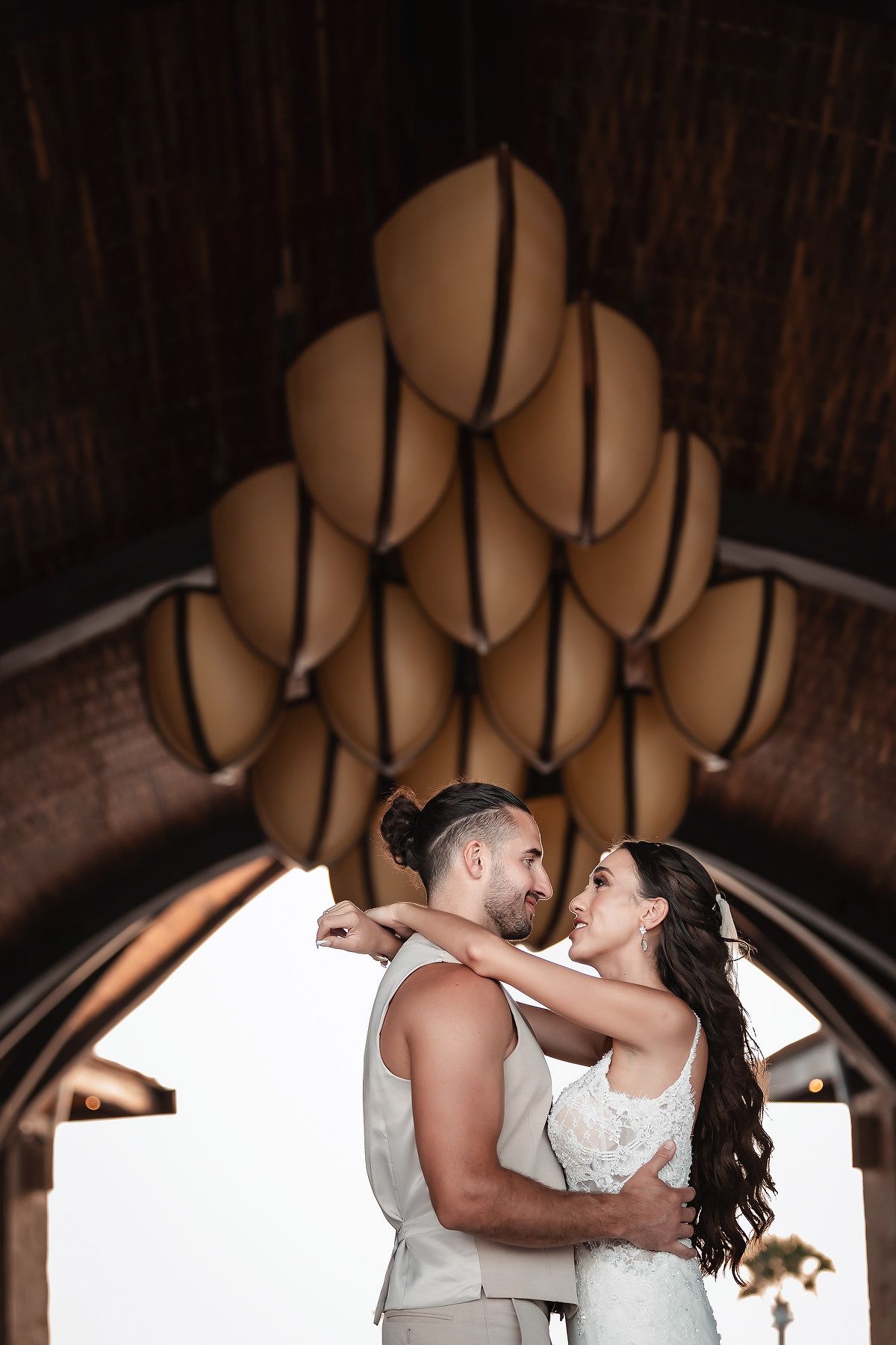 Couple embracing under decorative chandelier at wedding venue with wooden ceiling architecture.