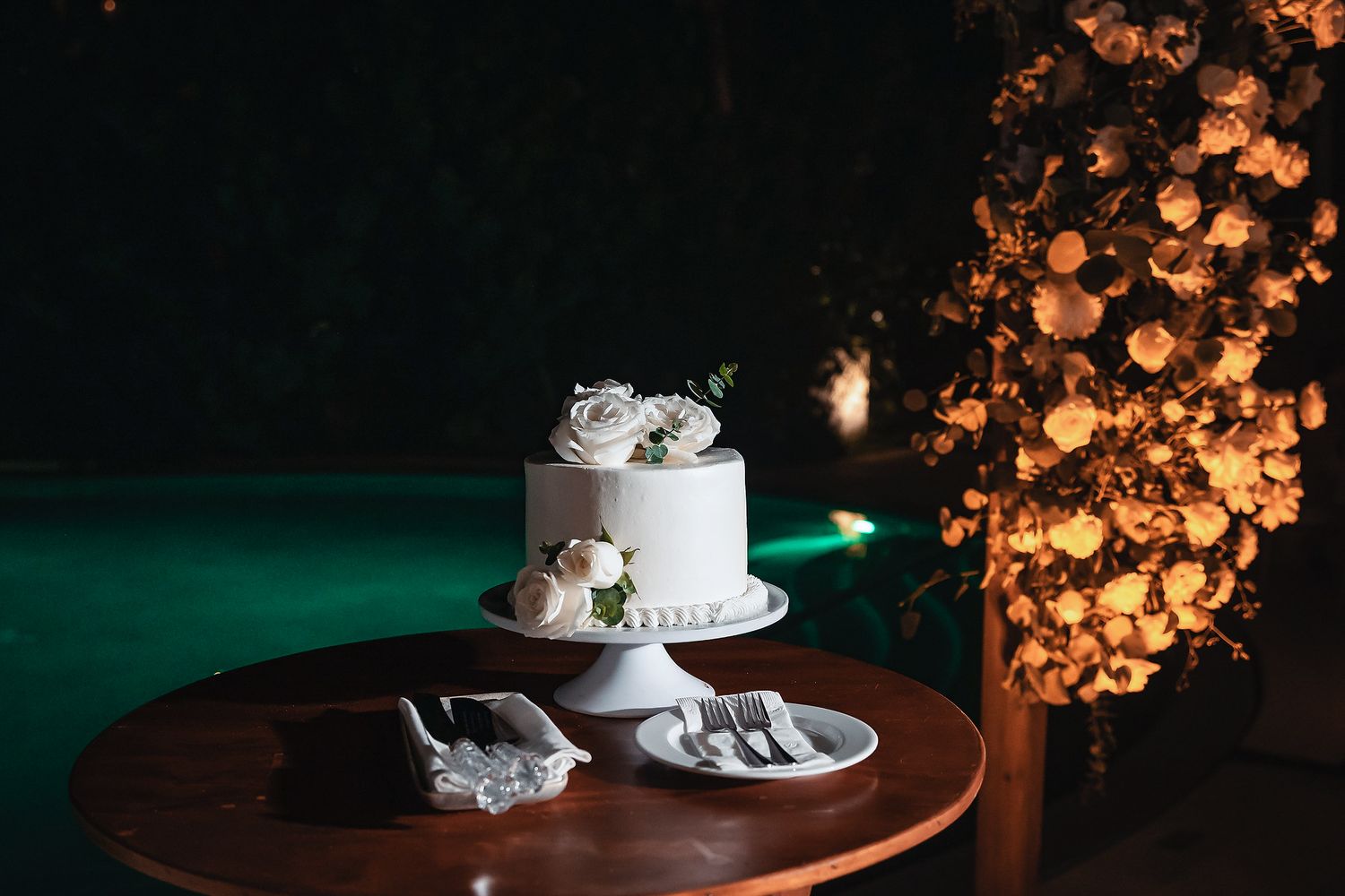 Elegant single-tier white wedding cake with floral topping on wooden table, beside glowing flower wall near pool at night.