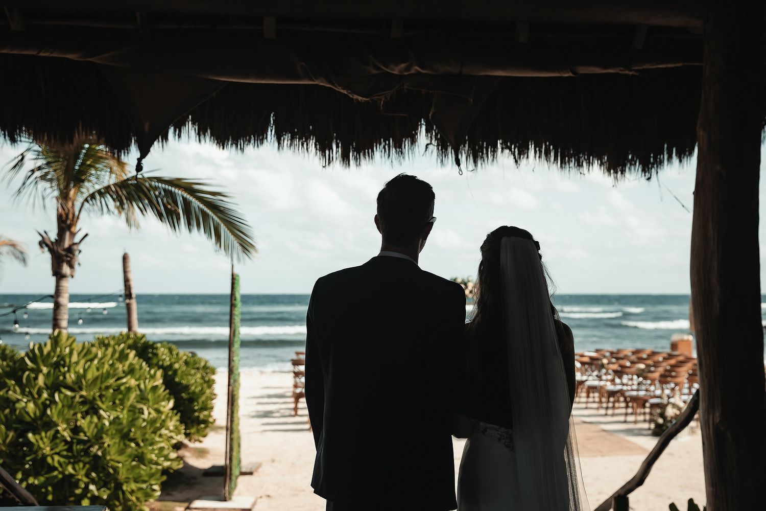 Silhouette of a bride and groom standing under a thatched roof gazebo overlooking a tropical beach on their wedding day.