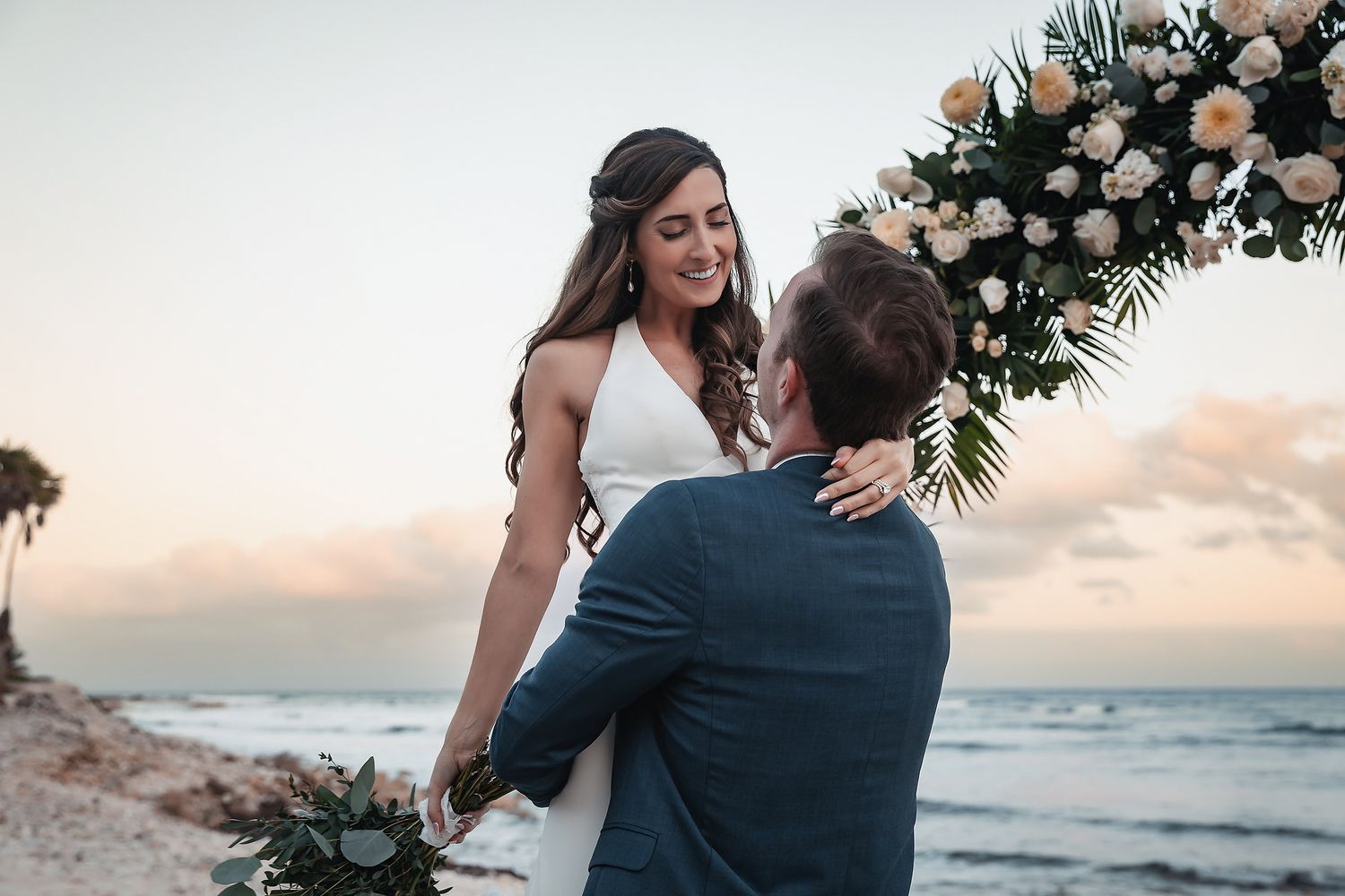 Bride and groom share romantic moment on beach at sunset near floral arch with white roses and tropical greenery.