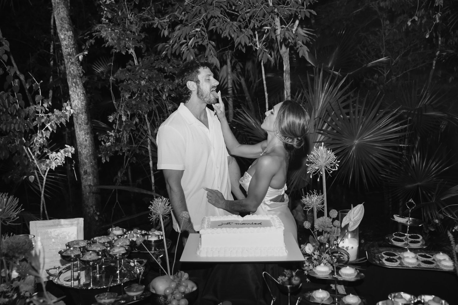 Black and white photo of newlyweds sharing cake at tropical outdoor wedding reception with floral dessert table.