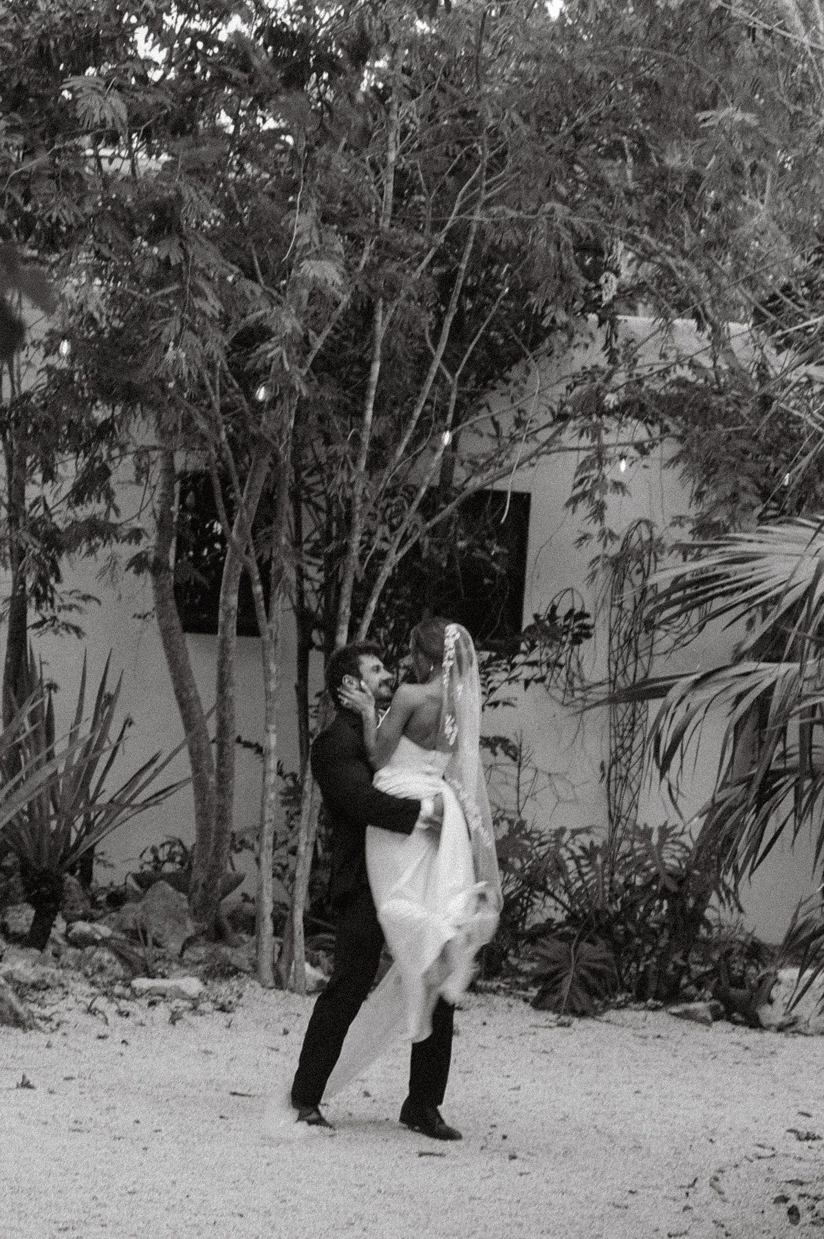 Groom lifting bride in white gown and veil outdoors among tropical trees in a romantic black and white wedding photo.