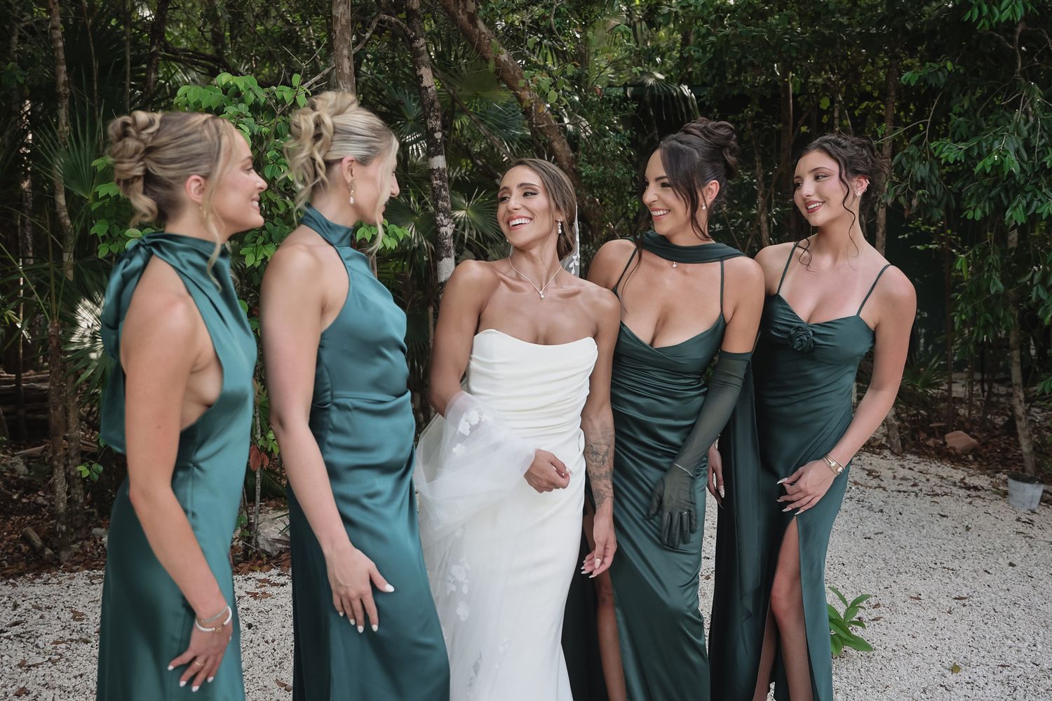 Bride in white strapless gown smiles with four bridesmaids in elegant green satin dresses outdoors.