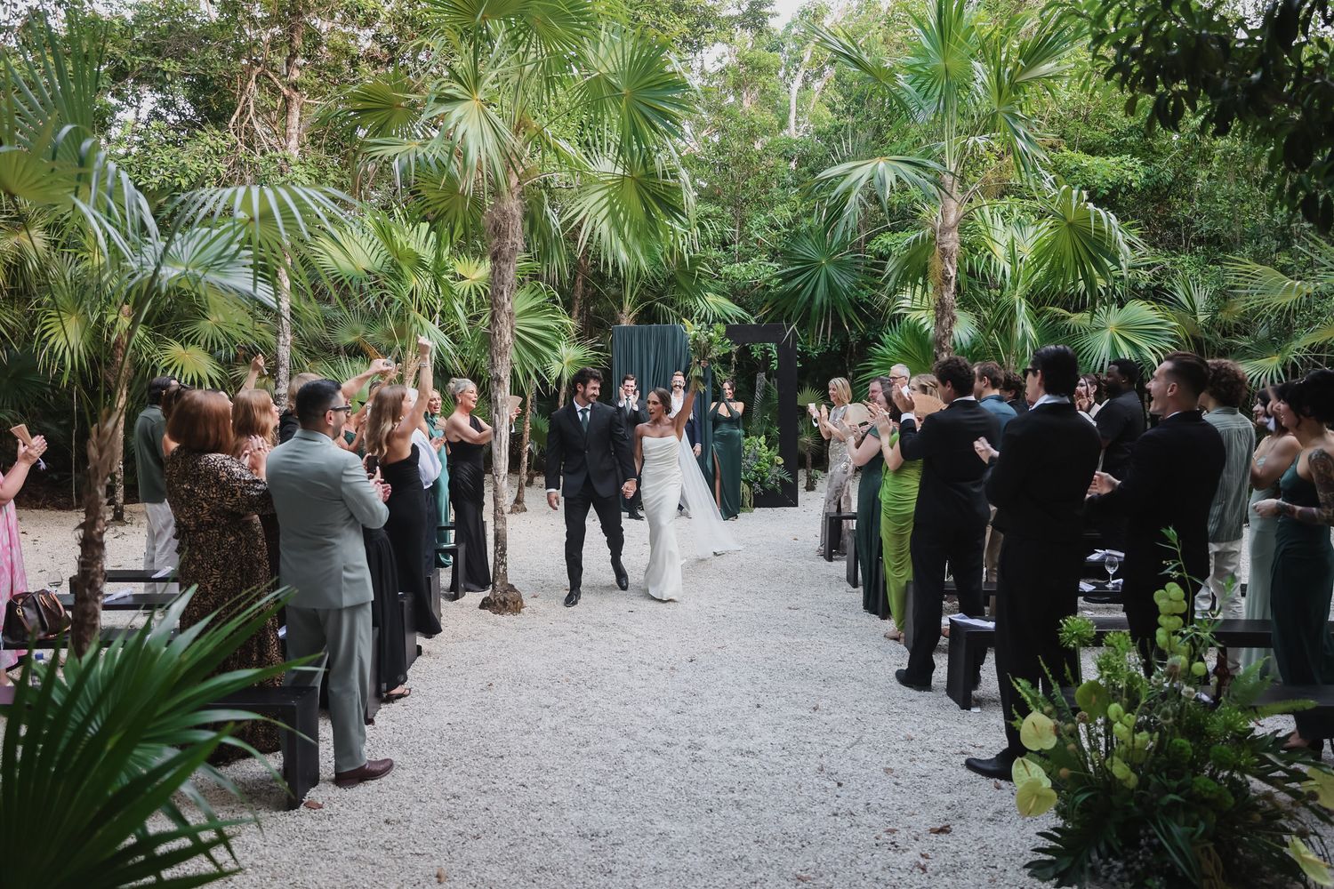 Bride and groom walk down outdoor aisle surrounded by lush palm trees and applauding guests at garden wedding.
