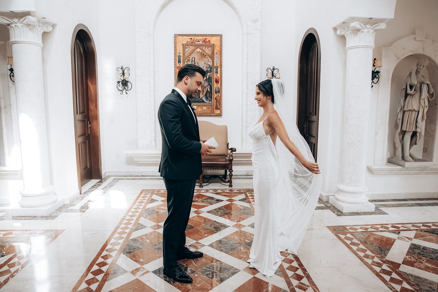 Wedding couple shares intimate first look moments in an elegant white marble hallway with ornate columns.
