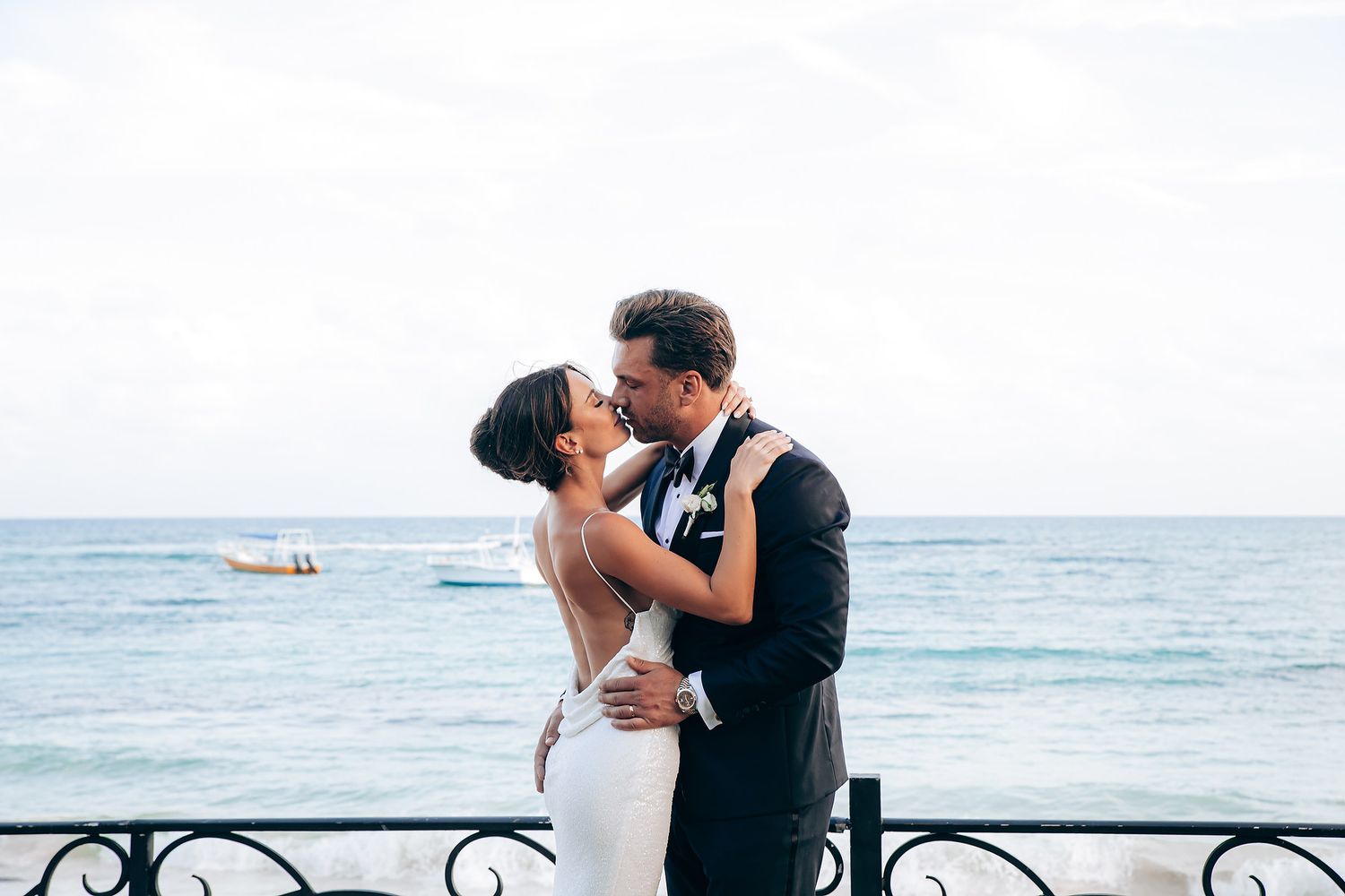 A romantic wedding couple embraces on a scenic beach with ocean waves and boats in the background.