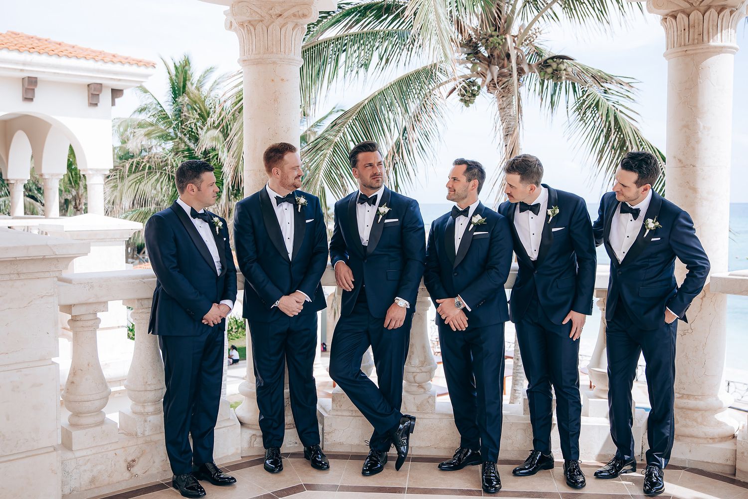 Groomsmen in navy suits lean against white stone pillars in a tropical outdoor setting during a wedding photoshoot.