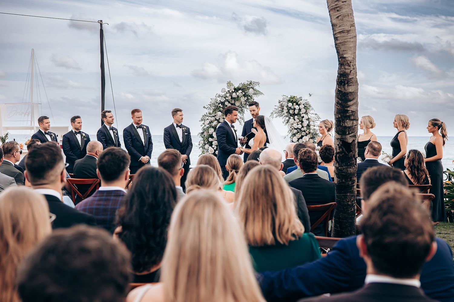 Wedding ceremony guests gather outdoors under cloudy skies with floral arch and ocean backdrop.
