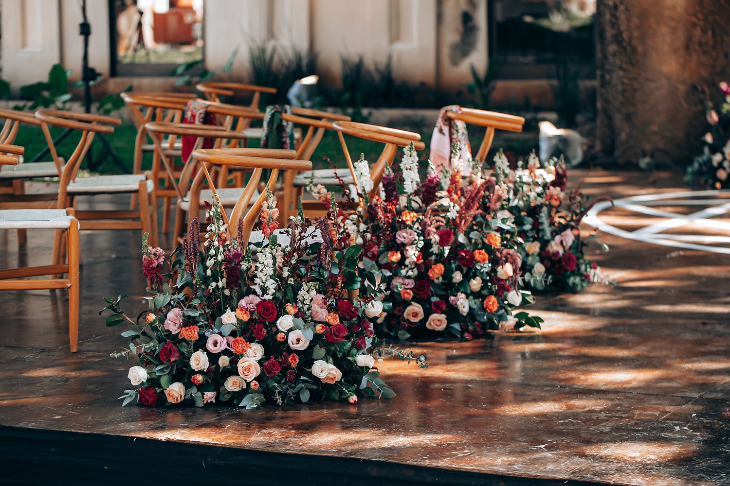 Wooden chairs and floral arrangements line an outdoor wedding ceremony aisle with pink and red blooms.
