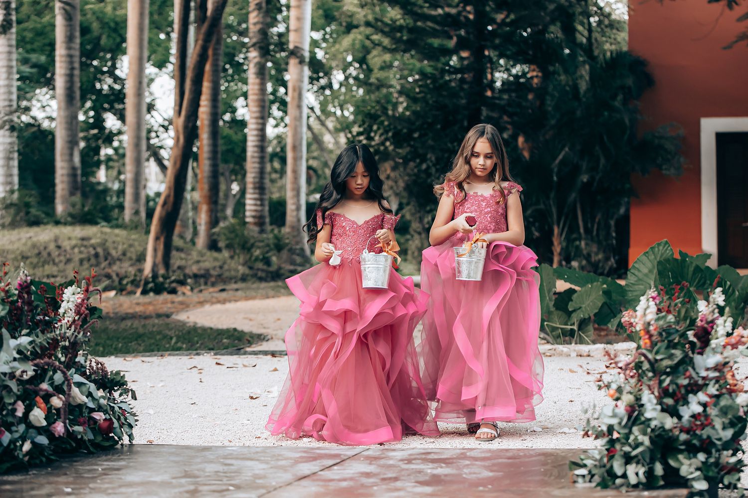Two young bridesmaids in matching pink dresses walk down a garden path carrying bouquets at an outdoor wedding.