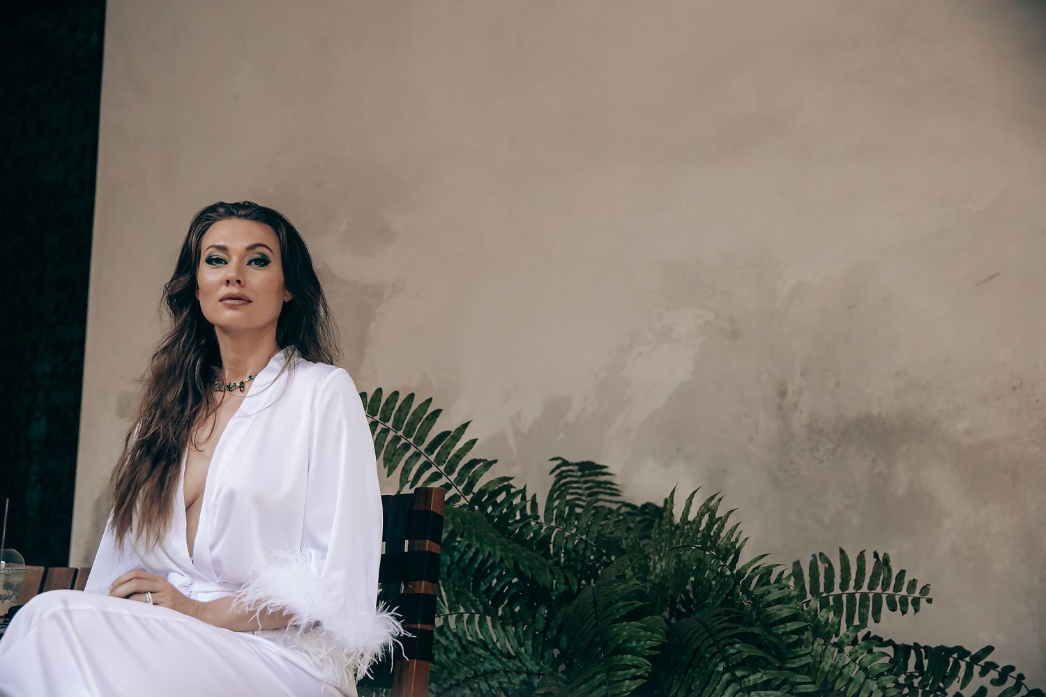 A person in a white outfit poses against a beige wall with green plants, creating an elegant outdoor portrait setting.