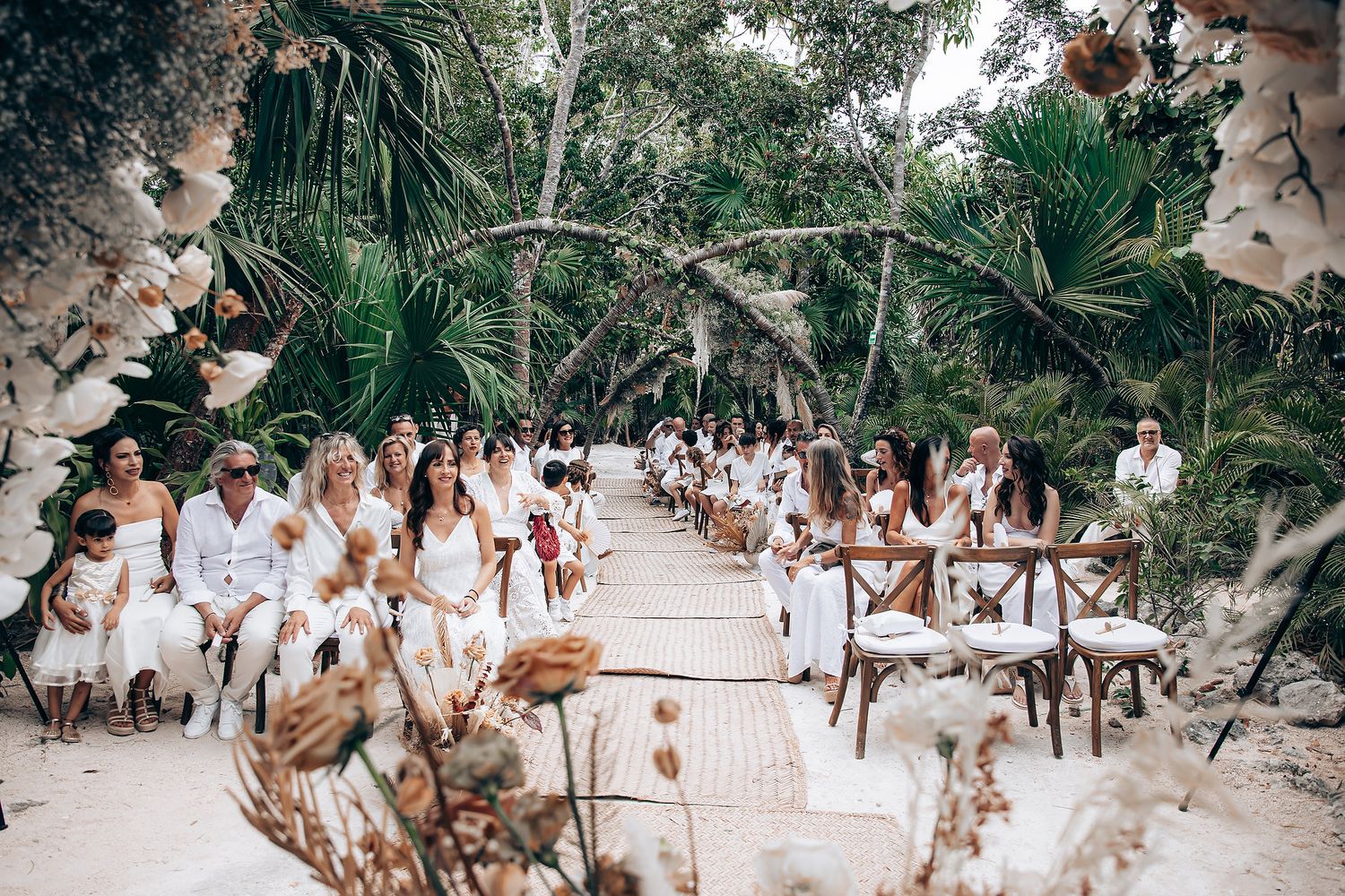 A tropical beach wedding ceremony setup with white chairs and floral arrangements along a sandy aisle.
