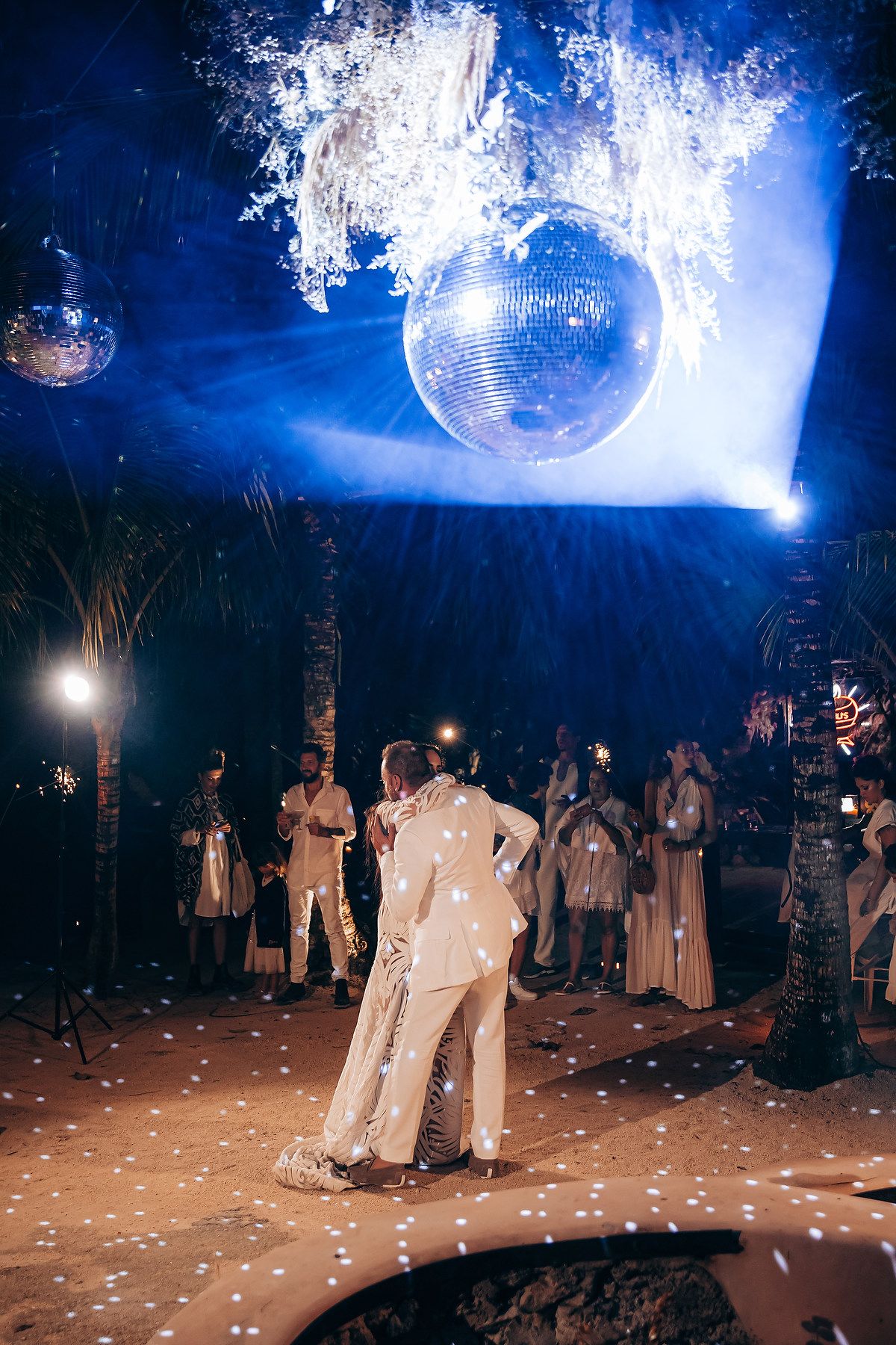 A couple dances under a sparkling disco ball and blue spotlights at an outdoor nighttime celebration.