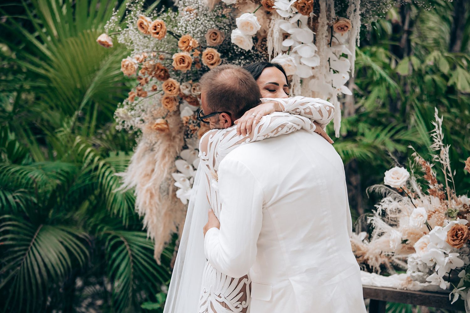 Couple embracing during wedding ceremony surrounded by lush tropical plants and peach floral arrangements.