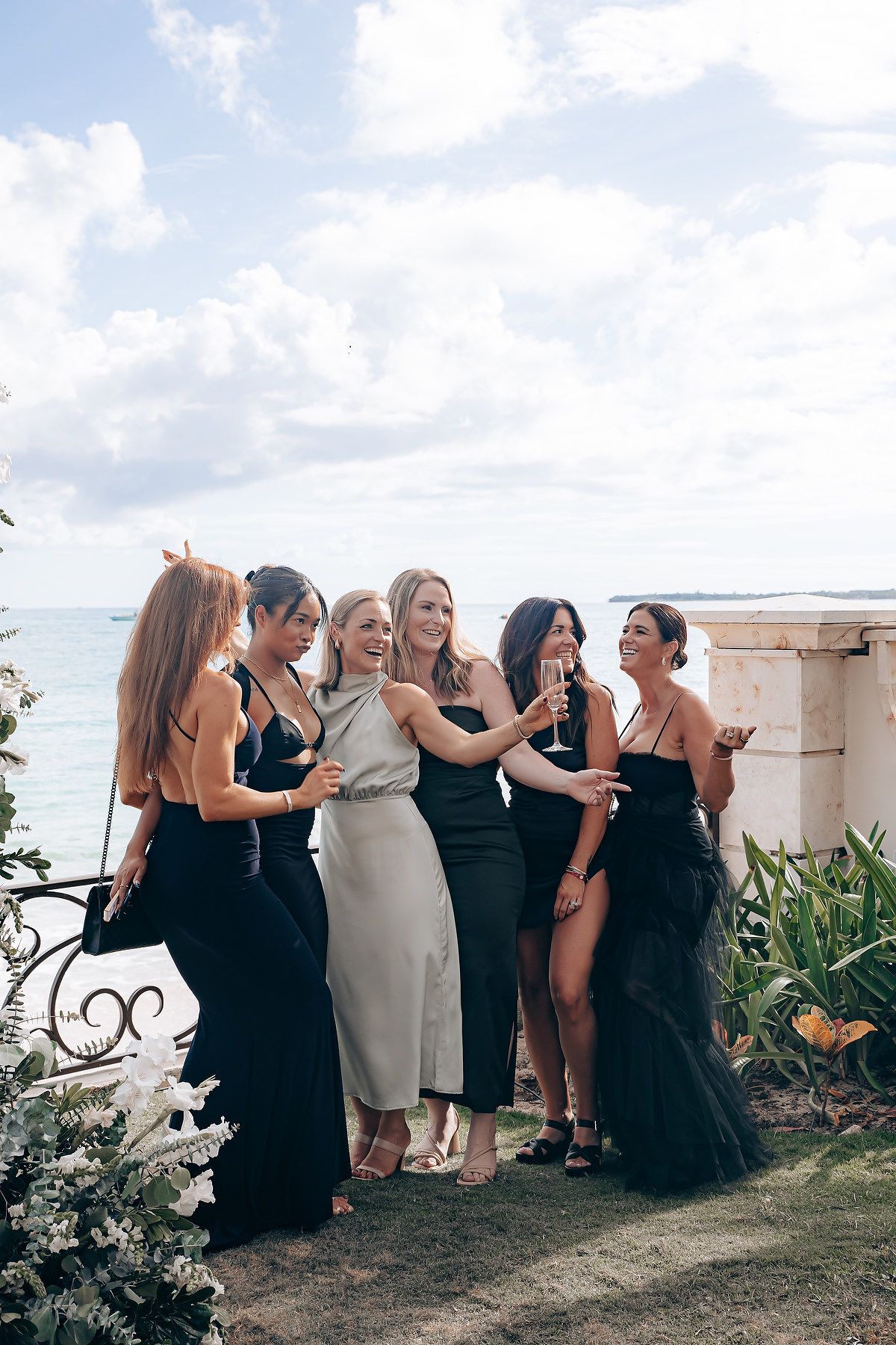 A group of women in elegant black and cream dresses laugh together at an outdoor wedding celebration by the ocean.