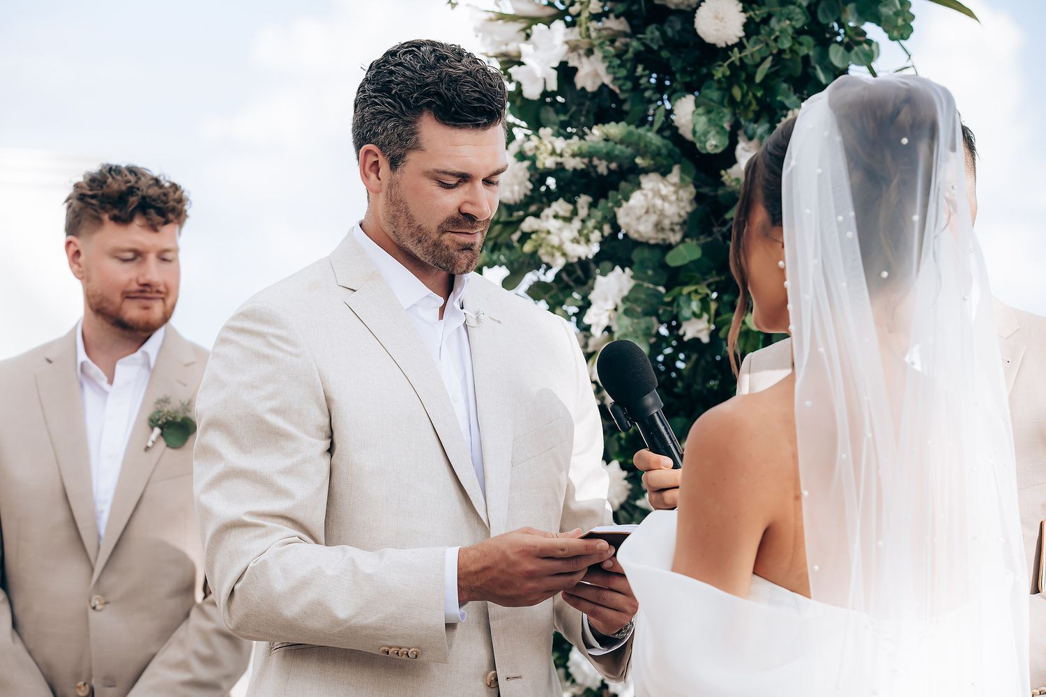 Wedding party members in light colored formal attire gather outdoors near green foliage during a ceremony.